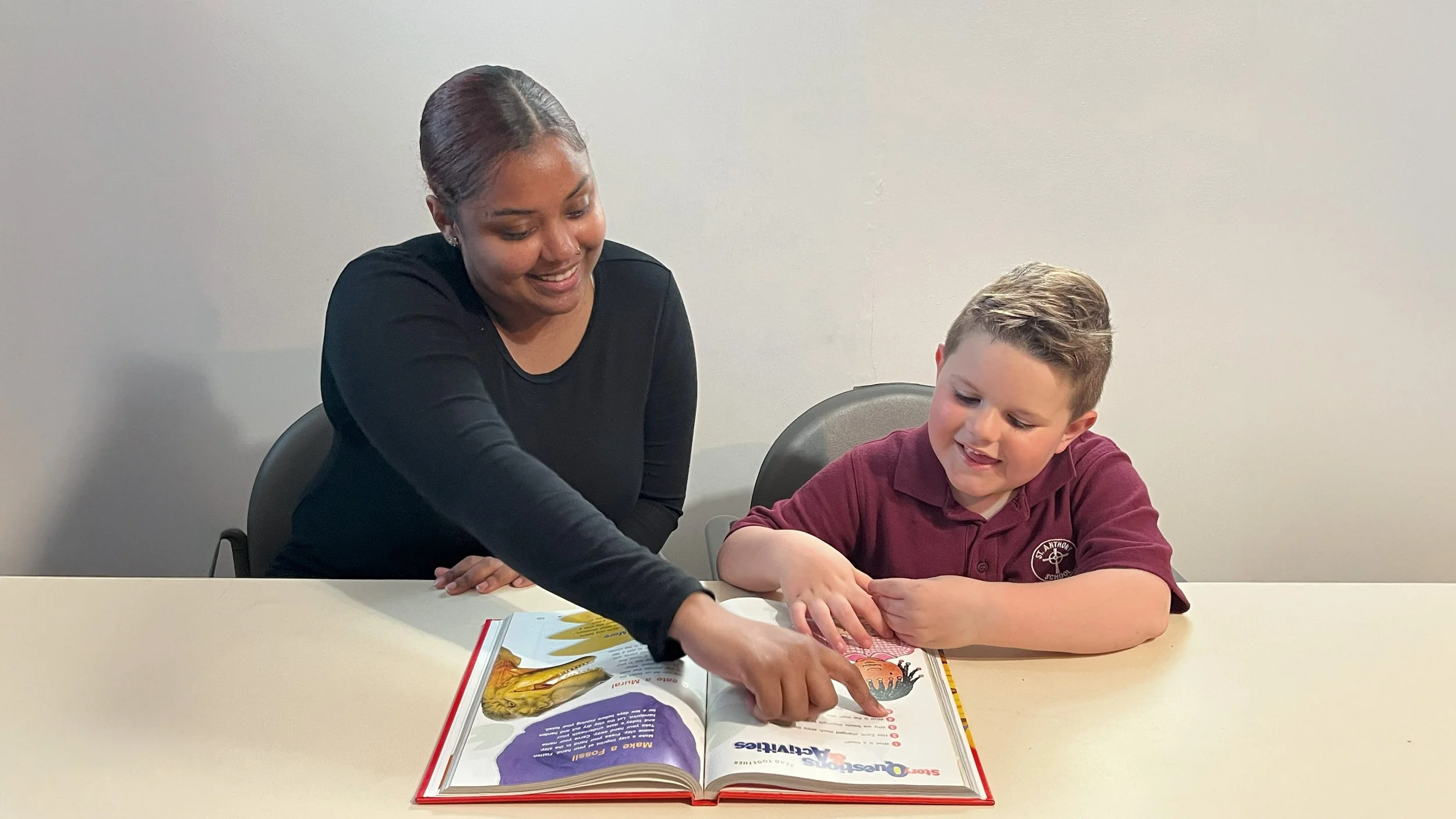 A woman and a young boy looking at a colorful book together at a table, smiling and engaging with the book.