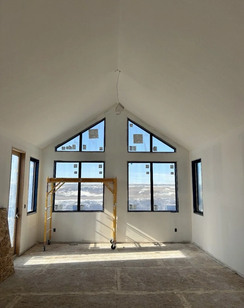Interior room under construction with large triangular and rectangular windows, unfinished flooring, and a yellow scaffolding in the center.