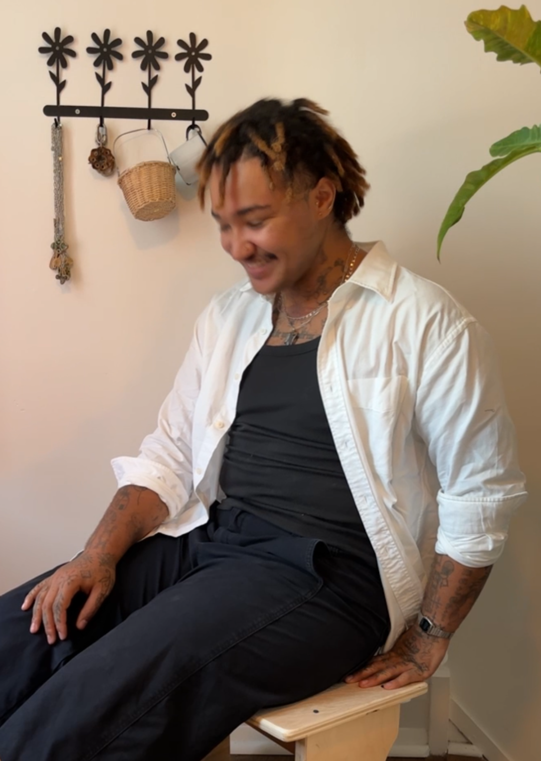A person with tattoos and dreadlocks, wearing a black shirt and a white button-up shirt, sitting on a wooden bench, smiling and looking down. A wall with decorative hooks, baskets, and hanging cups is visible in the background, along with some green leaves.