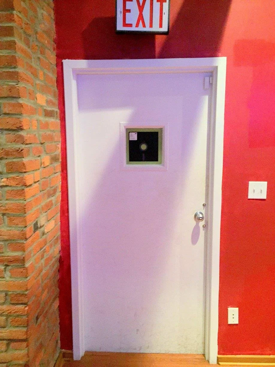White door with a small window in the upper center, situated in a red wall, with an exit sign above it. The door has a metallic doorknob and a push bar, opposite a brick wall.