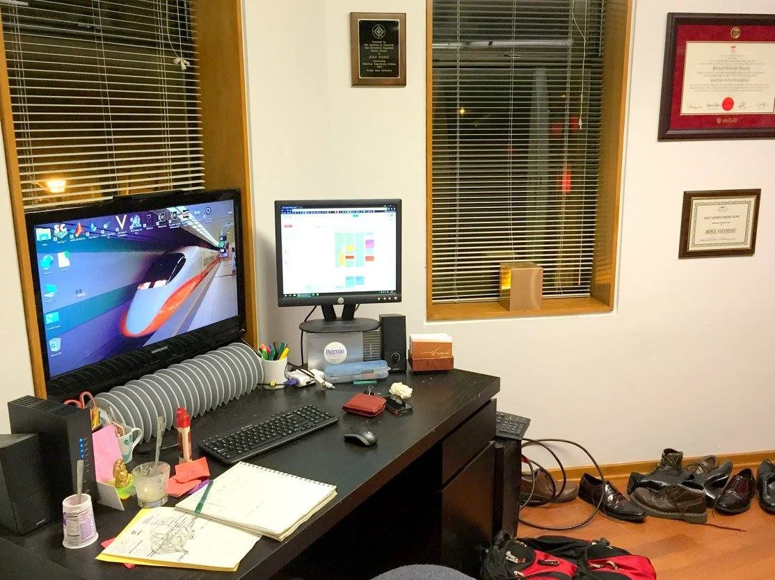 Office desk with two monitors, one showing a train wallpaper and the other a calendar or scheduling app. The desk has various office supplies, a notebook, and shoes lined up on the floor. There are framed certificates or awards on the wall and a wind