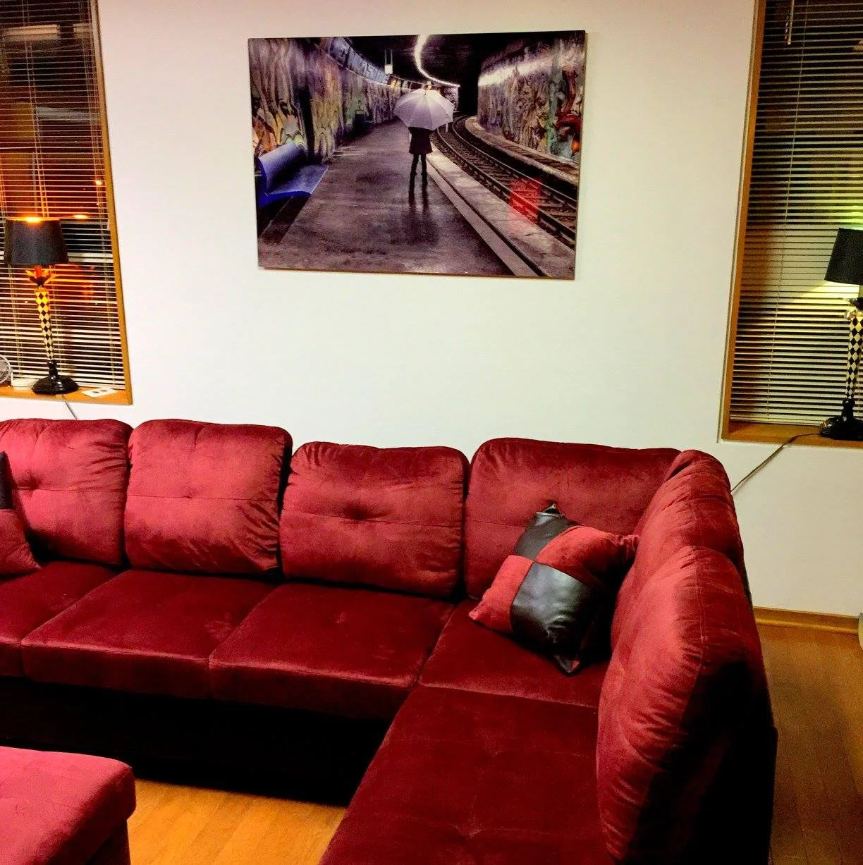 Living room with a red sectional sofa, two windows with blinds, a framed artwork of a person with an umbrella near a train track, and two black lamps on side tables.