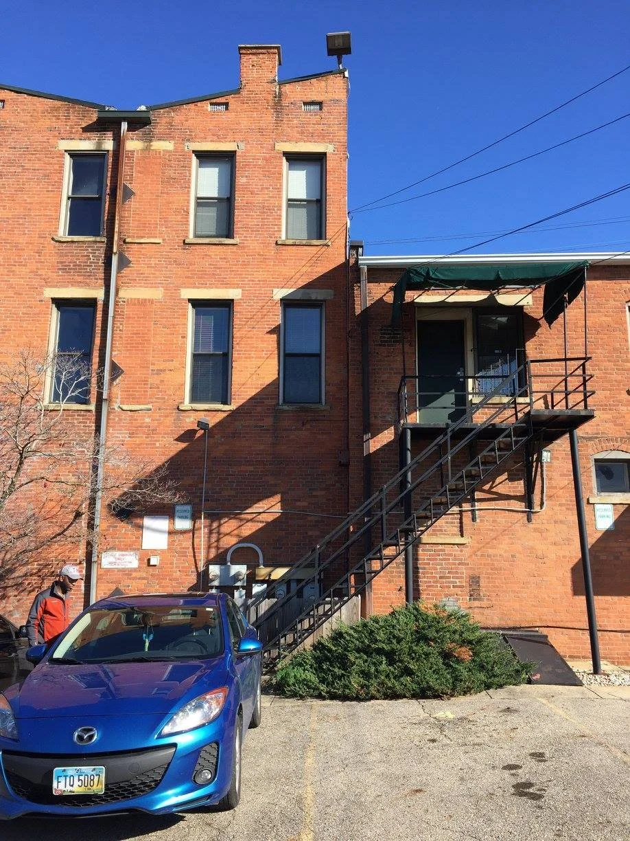 Red brick building with four windows, some with blinds, an external staircase with a green awning, a blue car parked in front, and a person in a red jacket and cap near the car under a clear blue sky.
