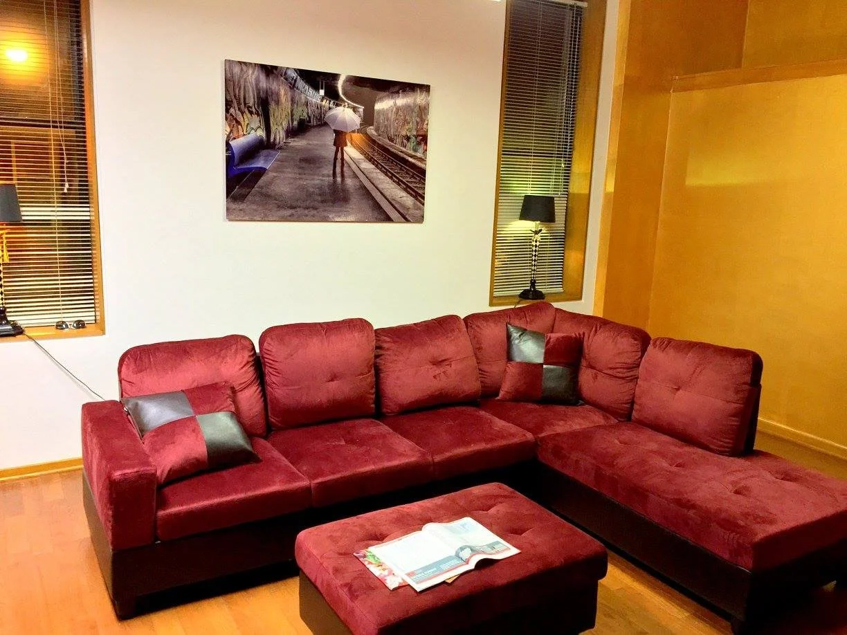 Living room with red sectional sofa, black and red throw pillows, wooden floor, white and yellow walls, a framed photo on the wall, two wall lamps, and windows with blinds.
