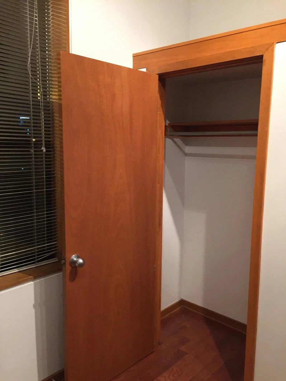 Empty wooden closet with an open door, located next to a window with blinds, inside a room with hardwood floor and white walls.