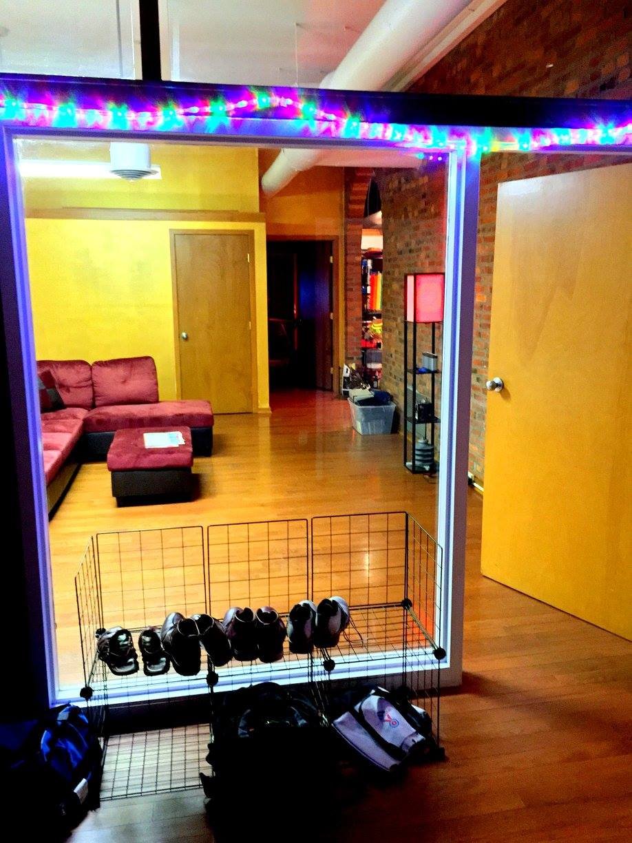 A storage cage with shoes and backpacks in front of a room with a yellow wall, red couch, and brick accents, illuminated by multicolored string lights.