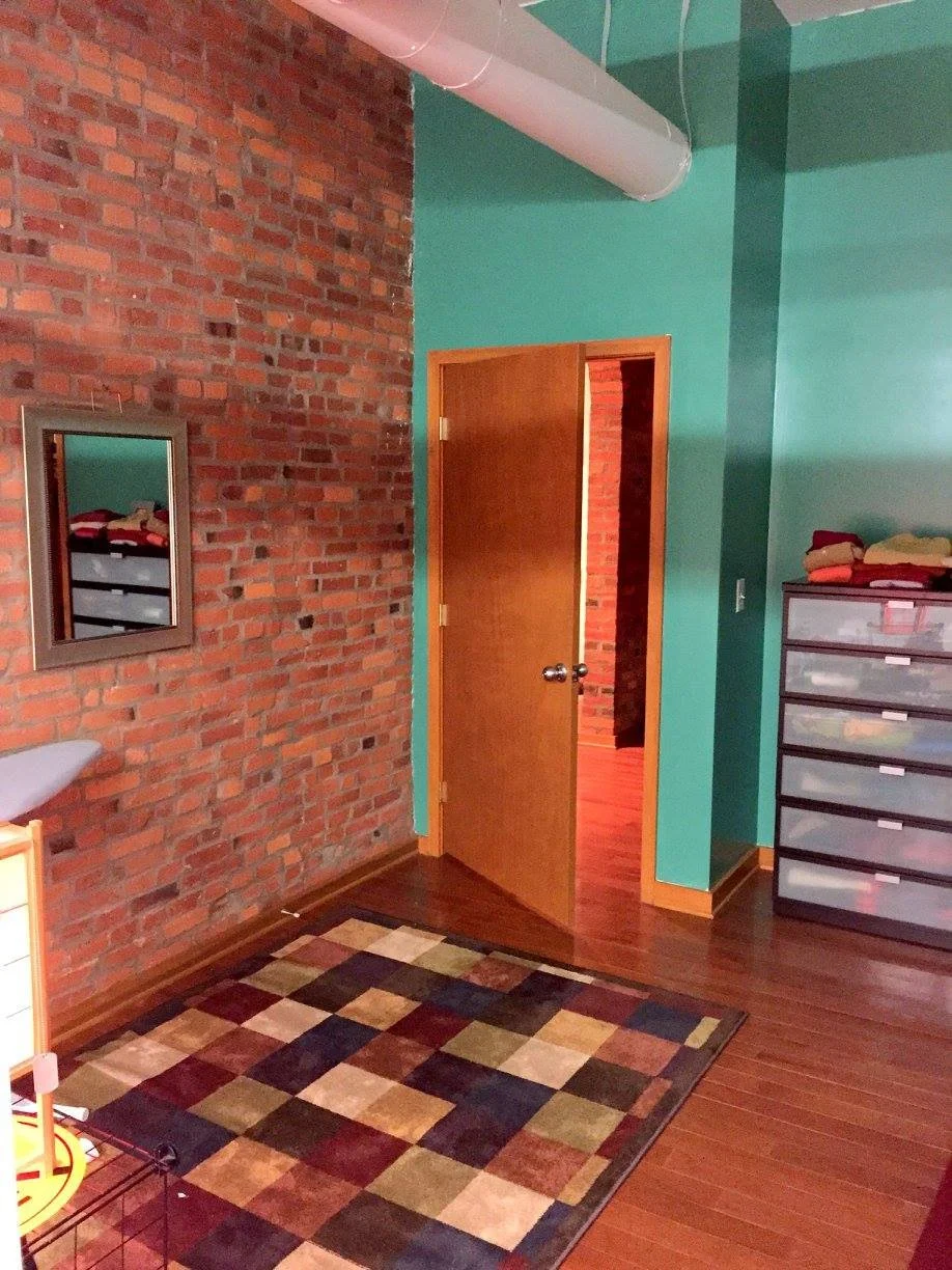 Interior view of a room with a brick wall on the left, turquoise painted walls, a wooden door slightly open, a colorful checkered rug on the wooden floor, and a plastic drawer unit with folded clothes on top.