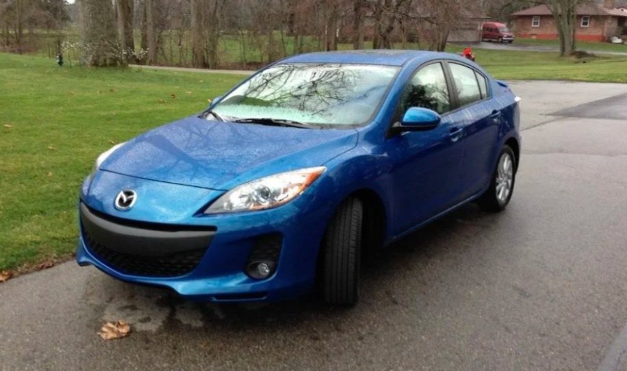 Blue Mazda sedan parked on the side of a neighborhood street on an overcast day with leafless trees and houses in the background.