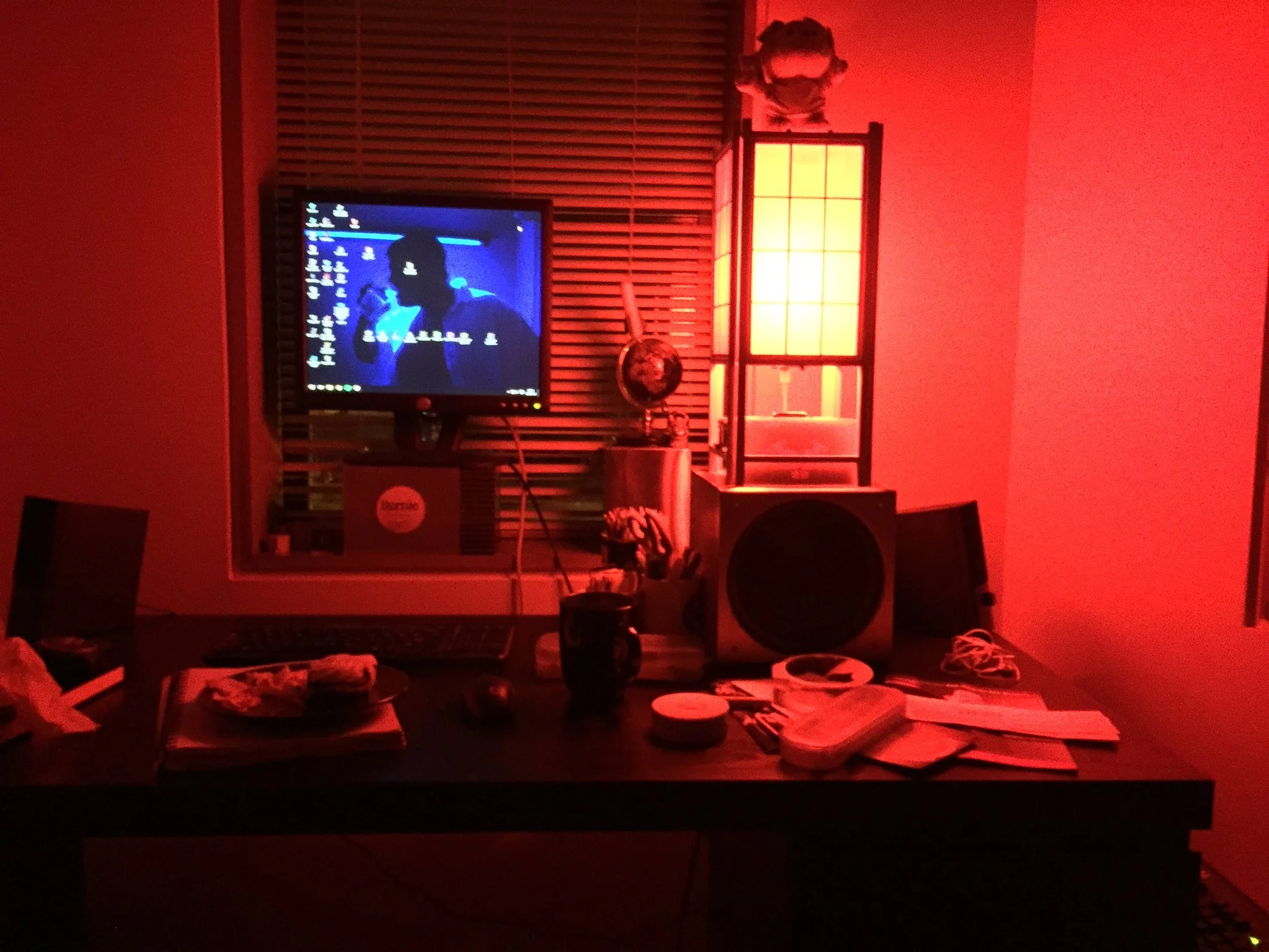 A dimly lit room illuminated by red light, with a computer monitor, scattered papers, and a window with blinds in the background.