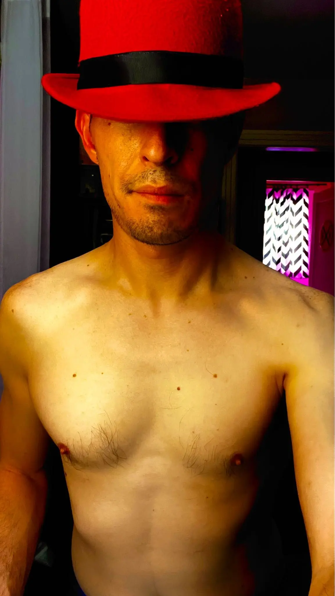 A person holding a smartphone, taking a selfie of themselves wearing a red hat with a black band, partially covering their face.