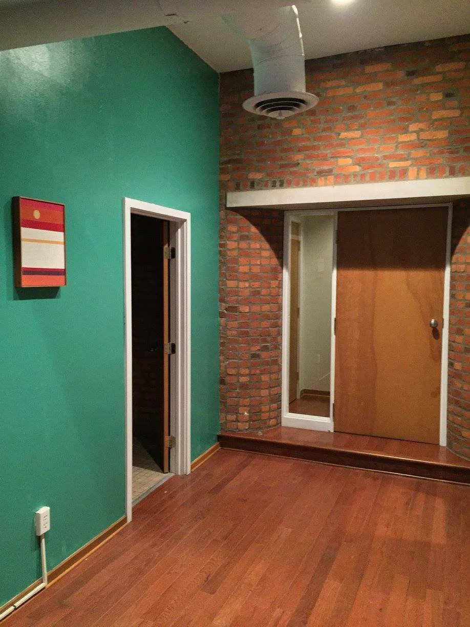 Room with green painted wall, brick wall, hardwood floors, a small wall mirror, a door, and a ceiling vent.