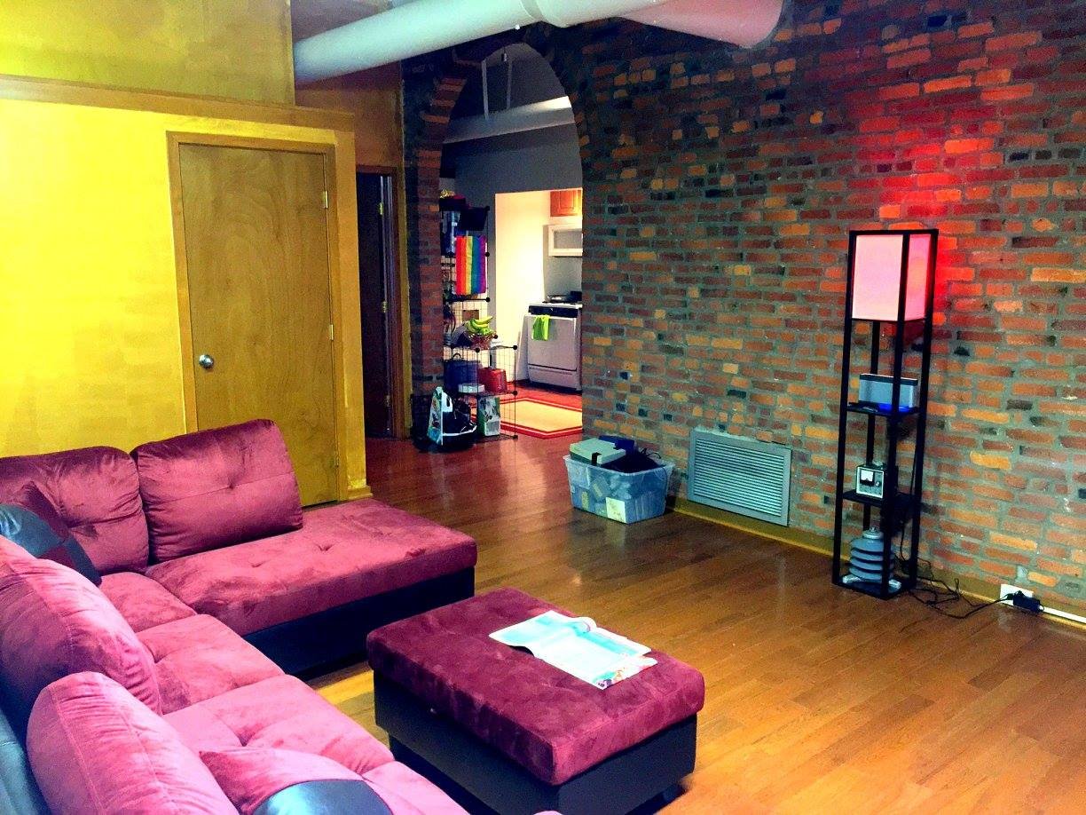 Living room with a pink sectional sofa, a black ottoman, and a brick wall. In the background, there's a kitchen area with a white oven and colorful towels. The room has hardwood floors and a tall black shelf with a pink lamp, some books, and a small 