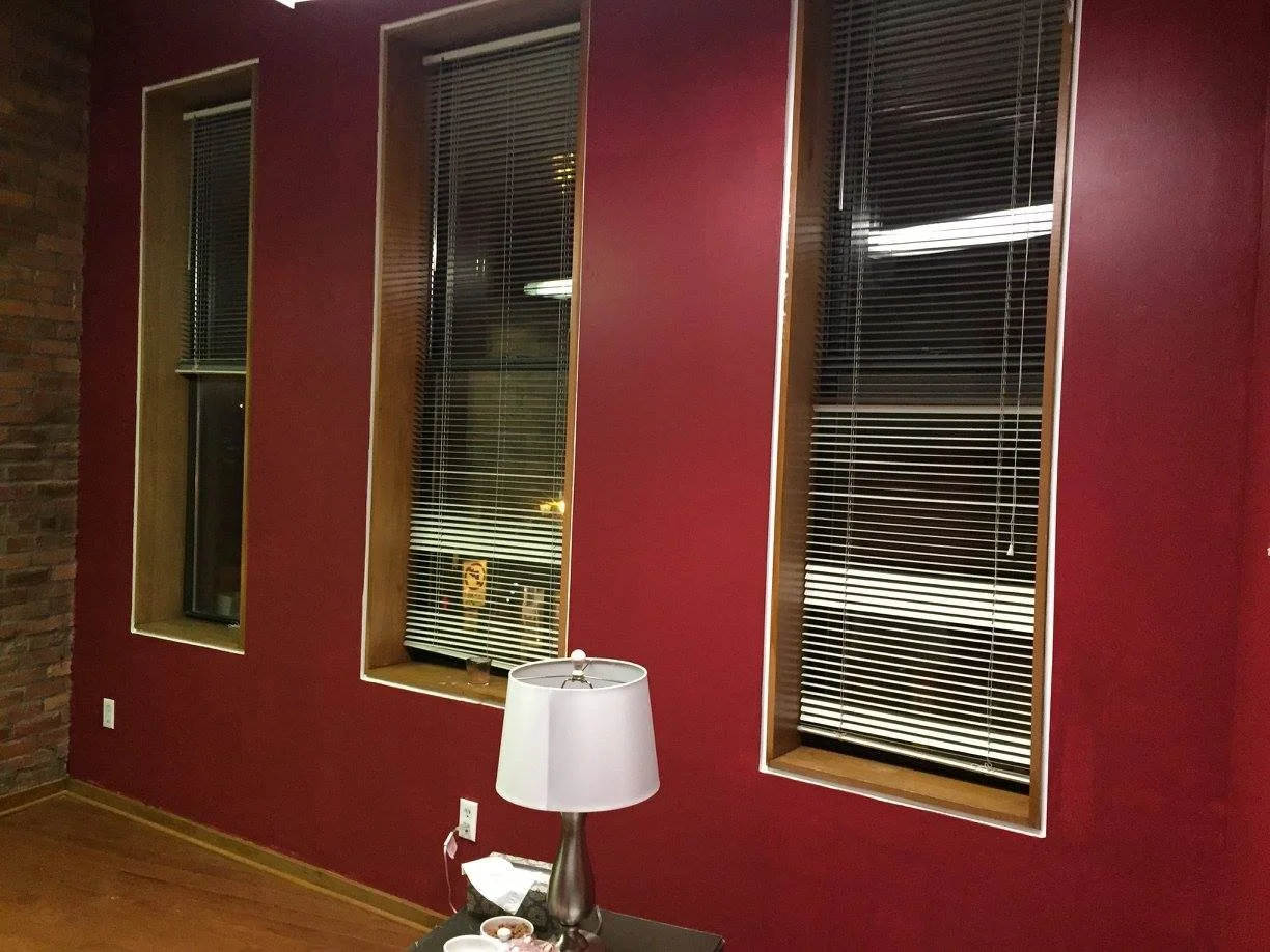 Interior of a room with a red wall and four large windows with blinds, a lamp on a table with some items, and a brick section on the left wall.