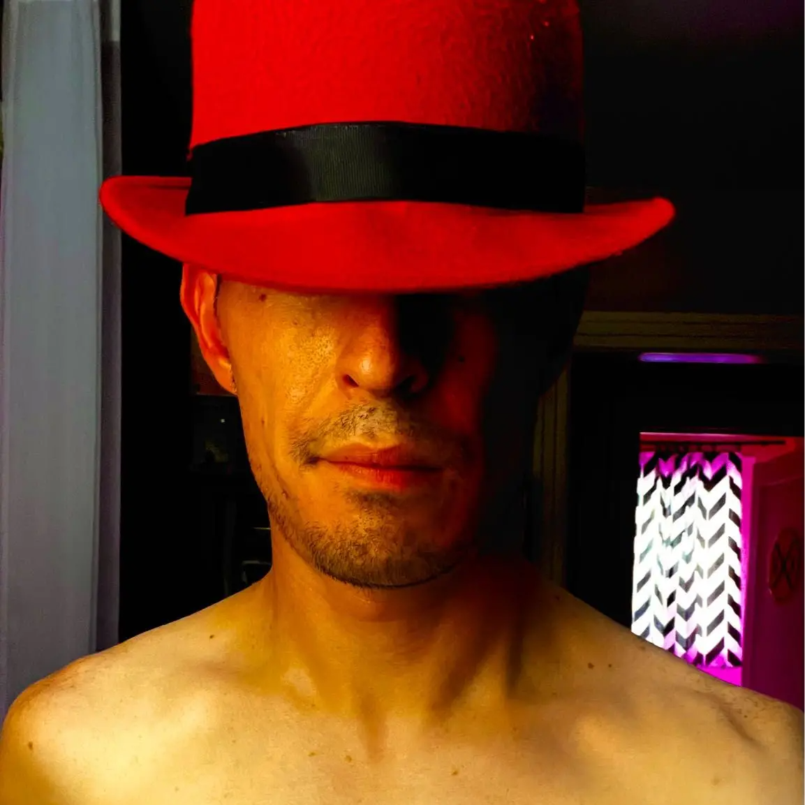 A person holding a smartphone, taking a selfie of themselves wearing a red hat with a black band, partially covering their face.