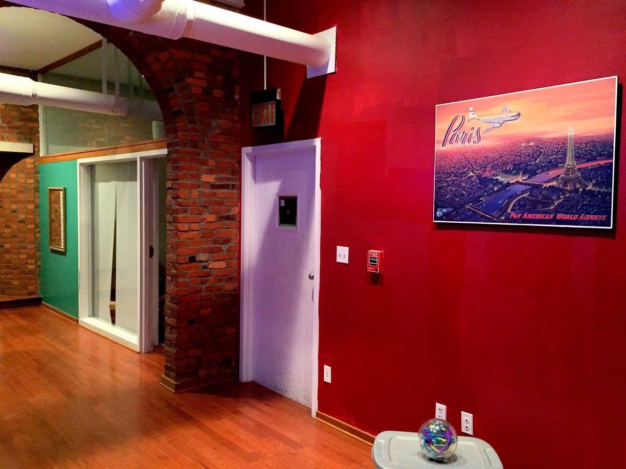 Interior of a room with a red wall, a framed Paris cityscape poster, a small white table with a decorative globe, brick archway, green accent wall, and a glass door.