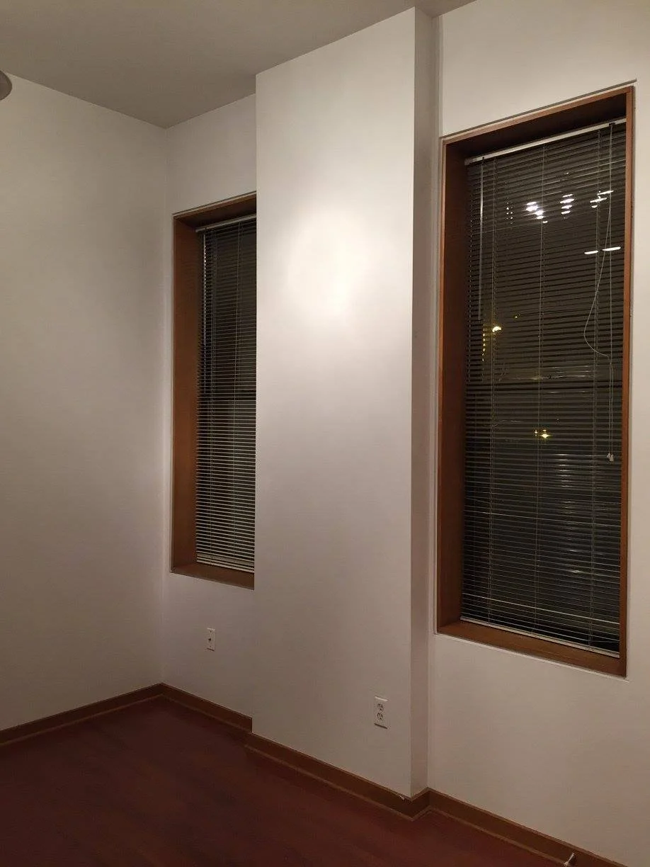 An empty room with two tall, narrow windows with closed blinds and wooden frames, white walls, and hardwood flooring.