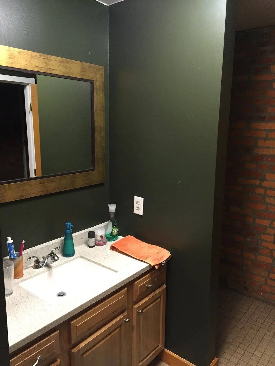 Bathroom with green walls, a mirror with a wooden frame, a beige countertop with a sink, a soap dispenser, a toothbrush holder, cleaning supplies, a folded orange towel, a wooden cabinet, tiled floor, and brick wall in the shower area.