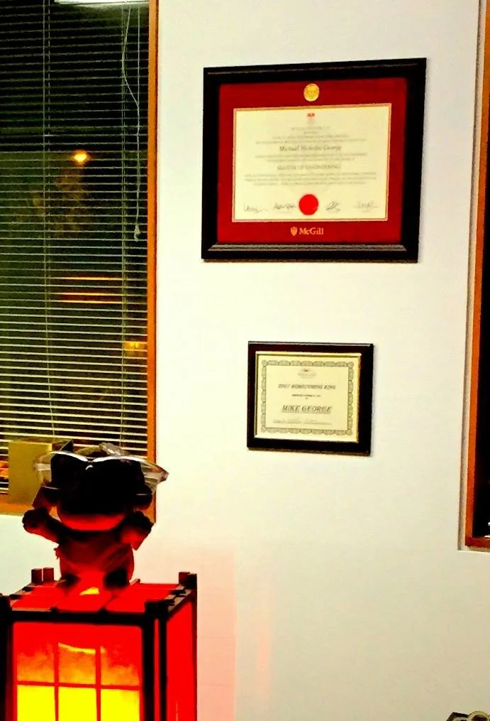 Wall with two framed certificates and a window with blinds. A plush toy of a red and black bear is placed on a red paper lantern with an orange glow.