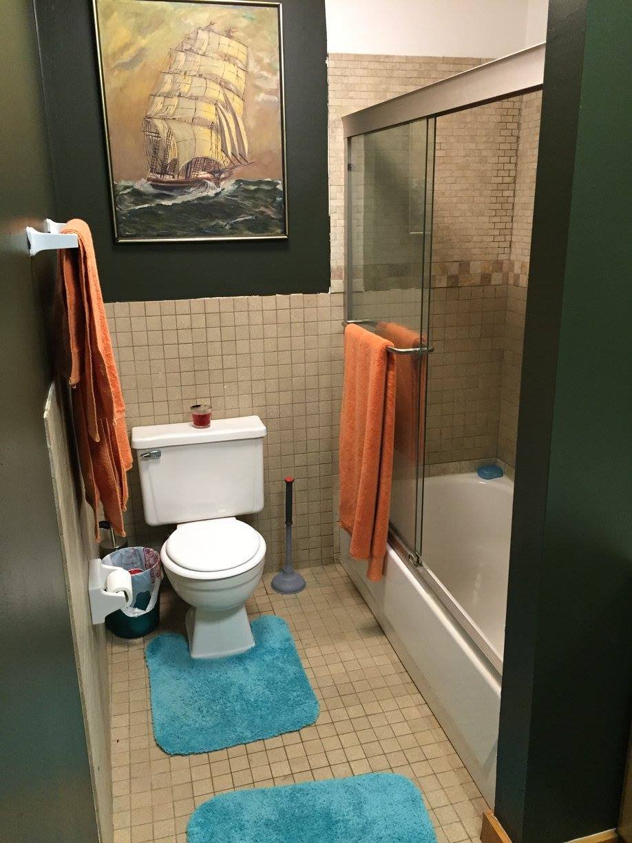 Small bathroom with beige tiled floor and walls, a white toilet with a teal rug in front, orange towels hanging on a rack and shower door, a framed sailing ship painting above the toilet, and a shower with glass sliding doors.