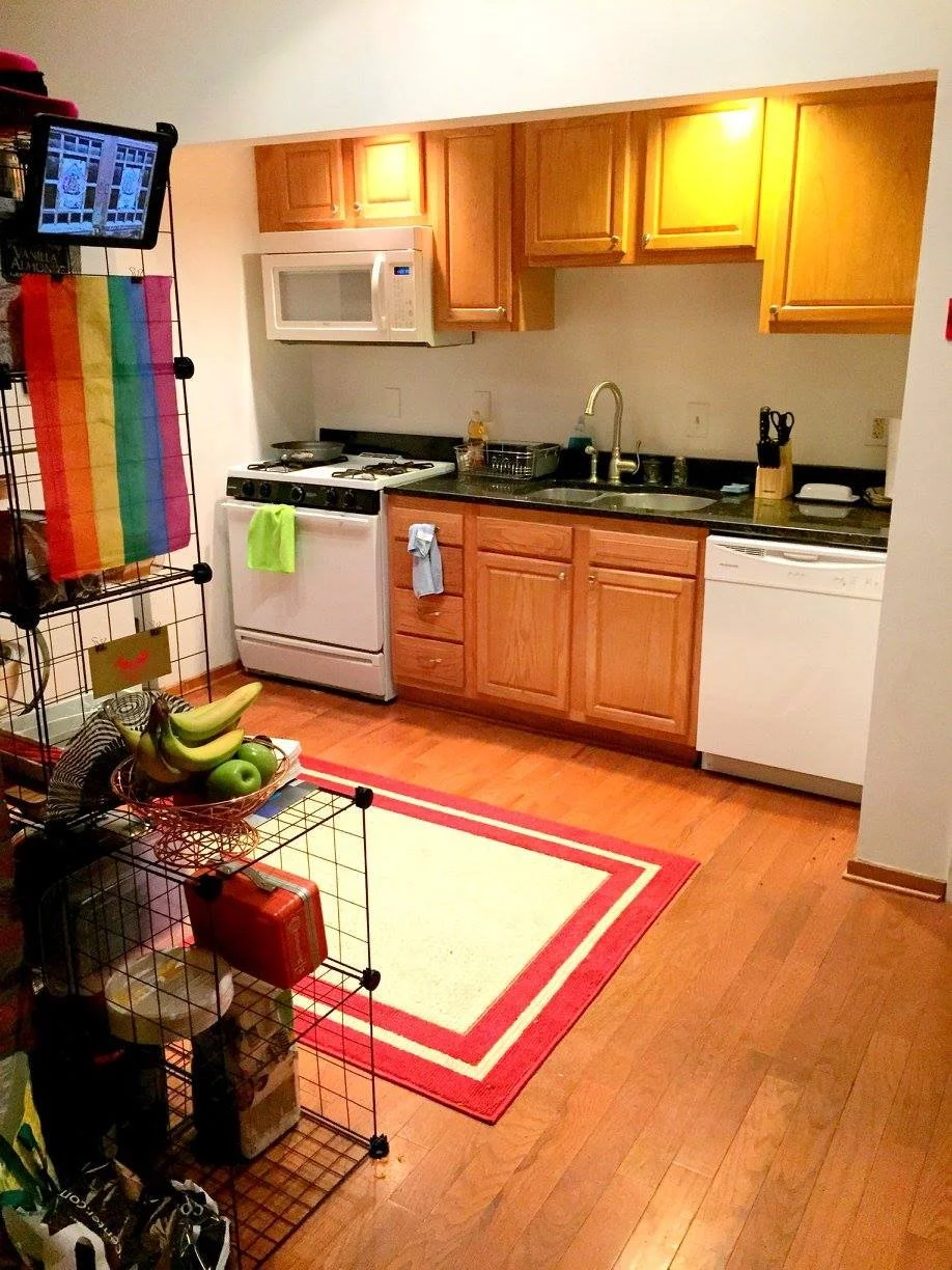 Small kitchen with wooden cabinets, black countertop, white microwave, stove, dishwasher, and a sink with a chrome faucet. There is a red and cream rug on wooden floor, and a black wire shelving unit with a fruit bowl and various items.