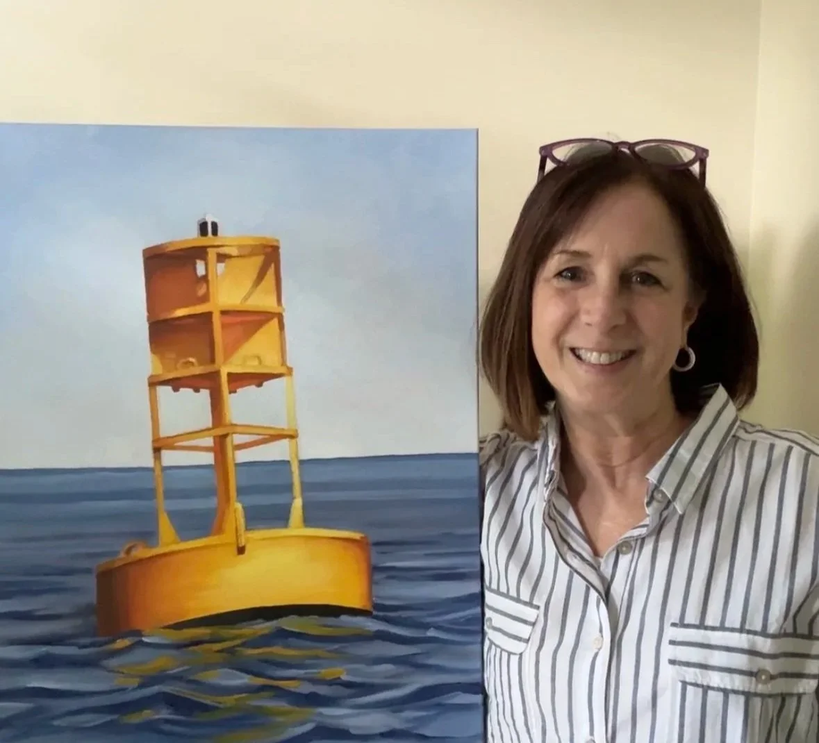Artist Tricia Granzier standing with glasses on her head smiling next to a painting of a bright yellow buoy in the water.