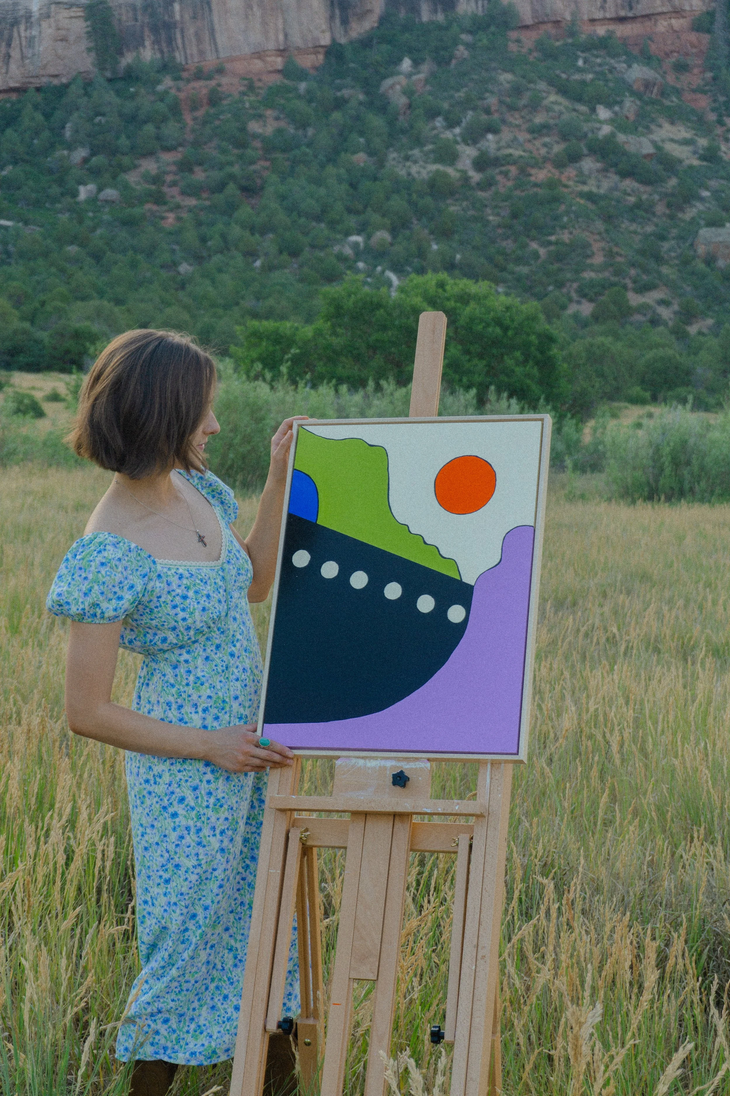 A woman in a floral dress paints a colorful abstract landscape on a wood panel outdoors, with a grassy field and mountains in the background during daytime.