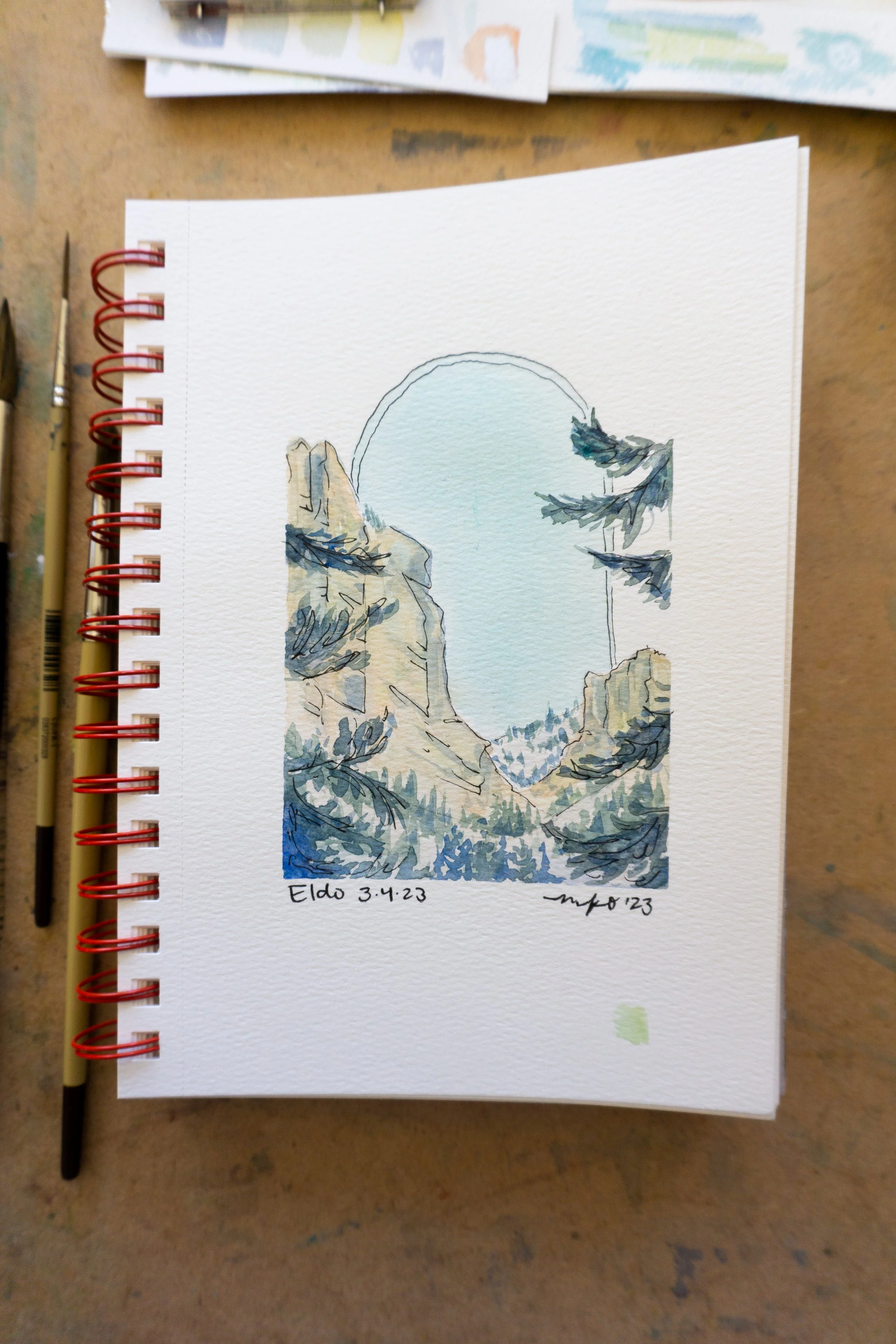 A watercolor painting of a mountainous landscape with trees, depicted in a sketchbook. The page is spiral-bound with a red wire. Paintbrushes are placed beside the sketchbook on a textured surface. The painting has an oval sky, rocky cliffs, and pine