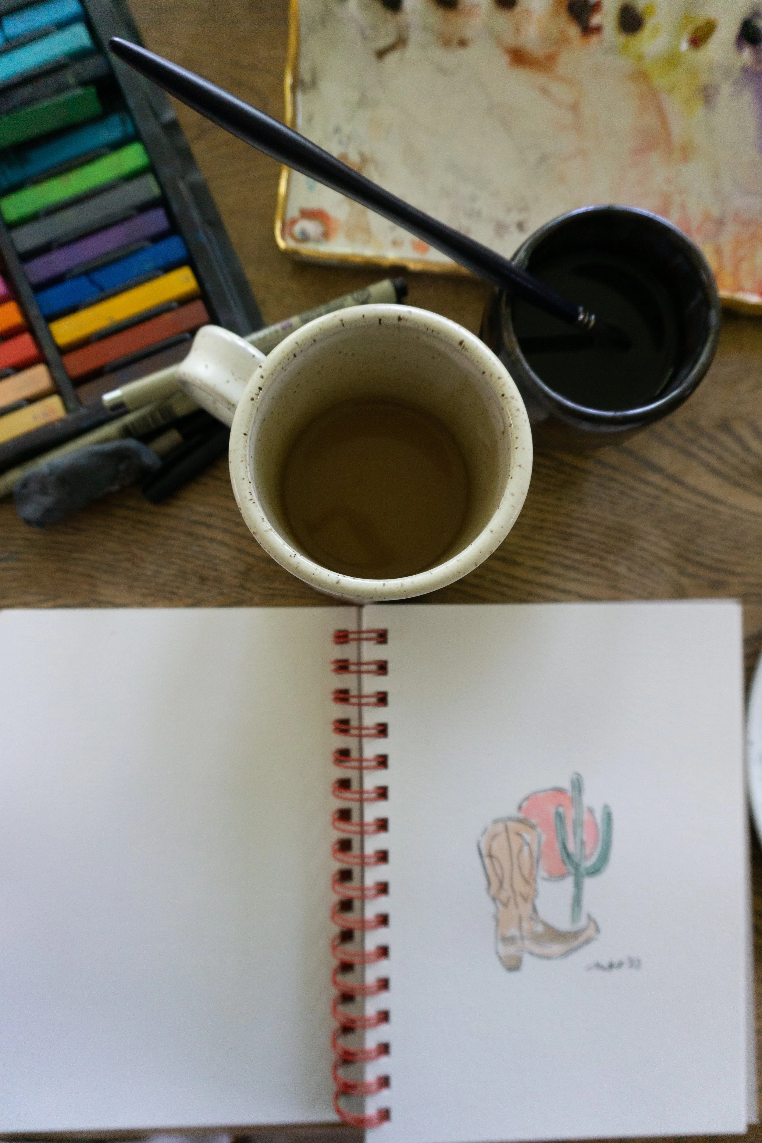 Art supplies including pastel sticks, a mug, brushes in water, and an open sketchbook with a drawing of a cowboy boot, cactus, and sun.