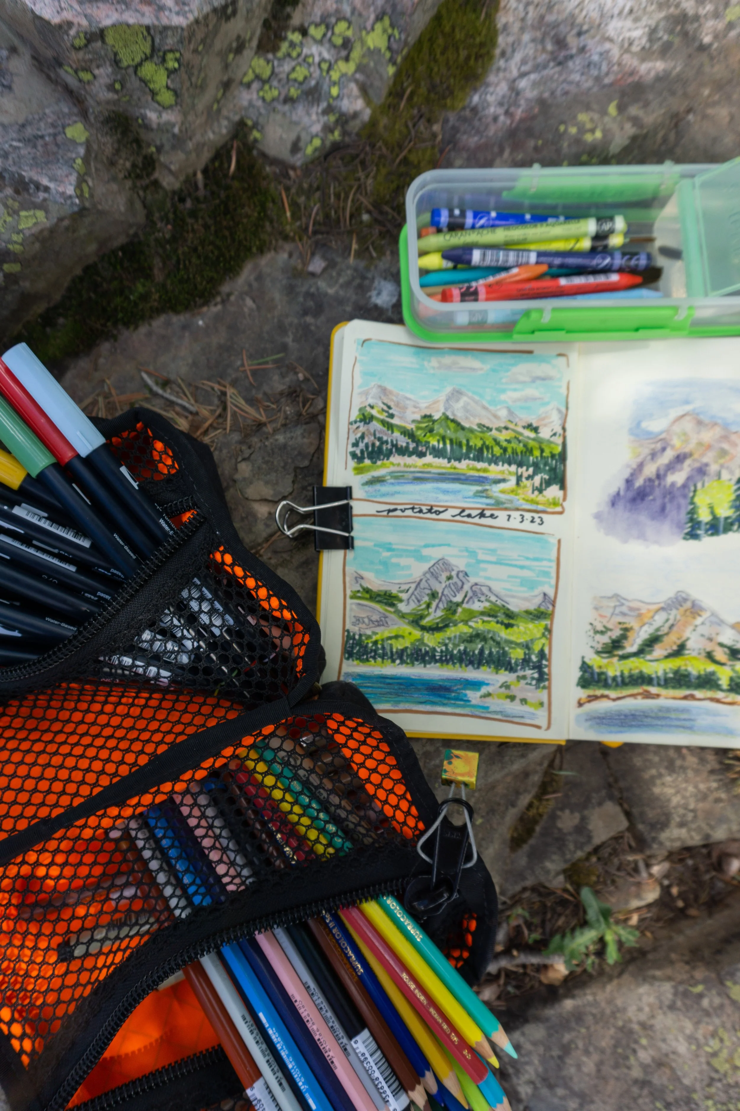 Art supplies on rocky ground, including colored pencils, markers, and a sketchbook with landscape paintings of mountains and a lake.