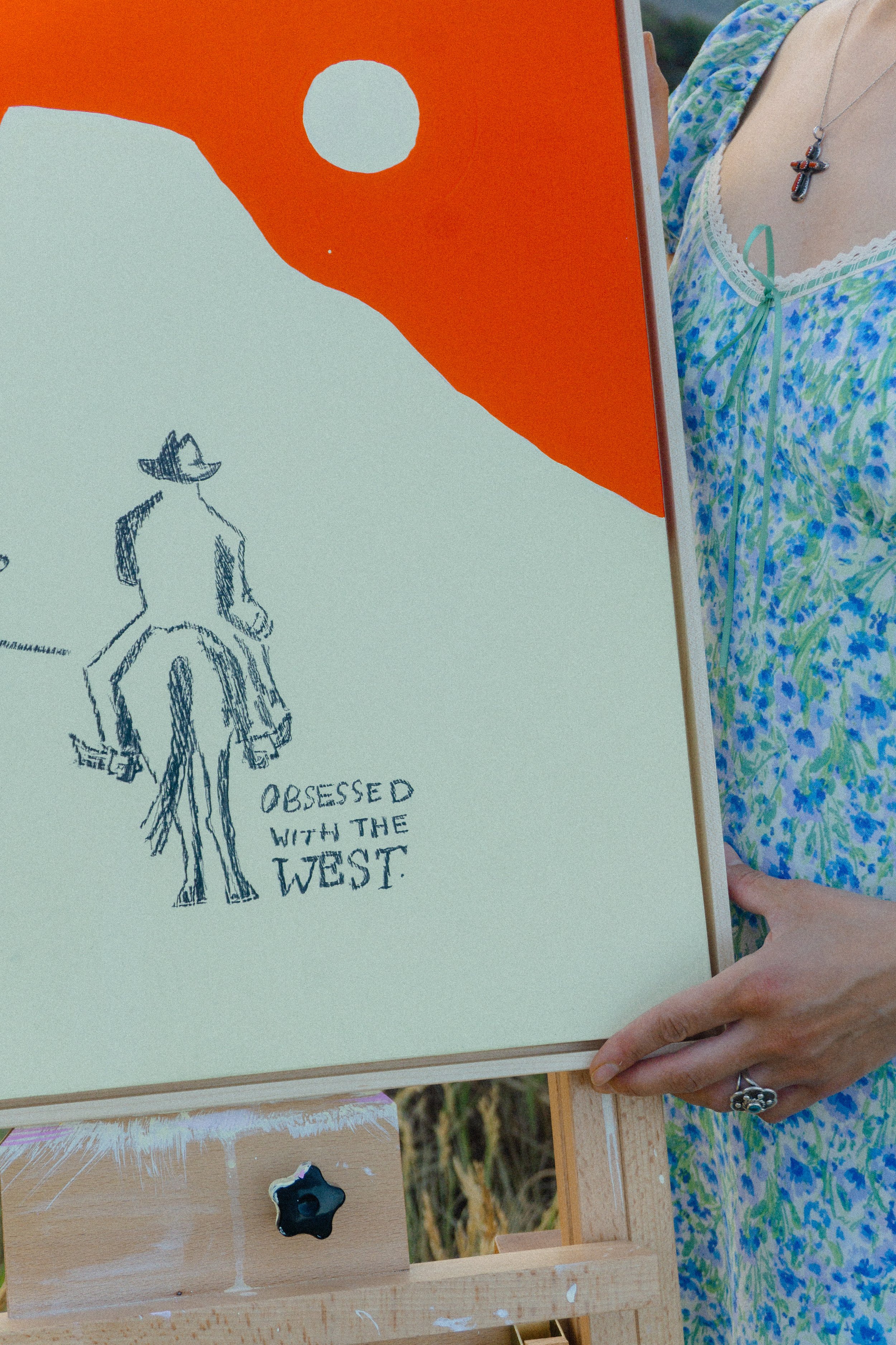 A person holding a sketchpad with a drawing of a cowboy riding a horse and the words "Obsessed with the West" underneath, with a painting of an red landscape and a white sun in the sky.