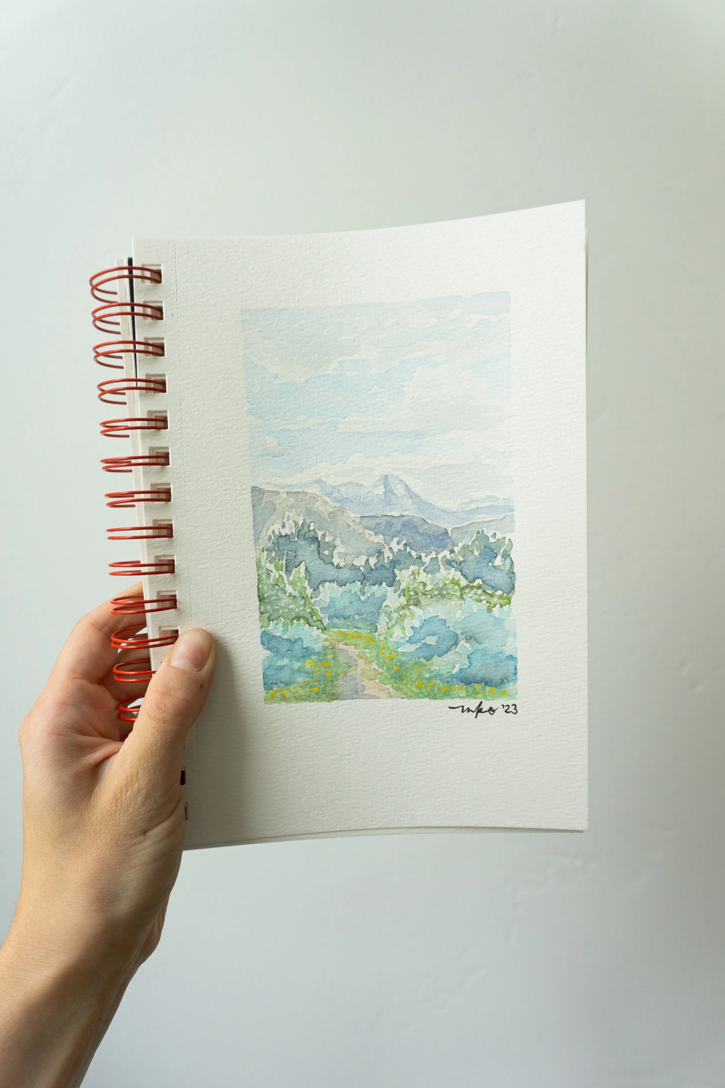 Hand holding a sketchbook with a watercolor painting of a landscape, featuring mountains, a blue sky, and green foliage.