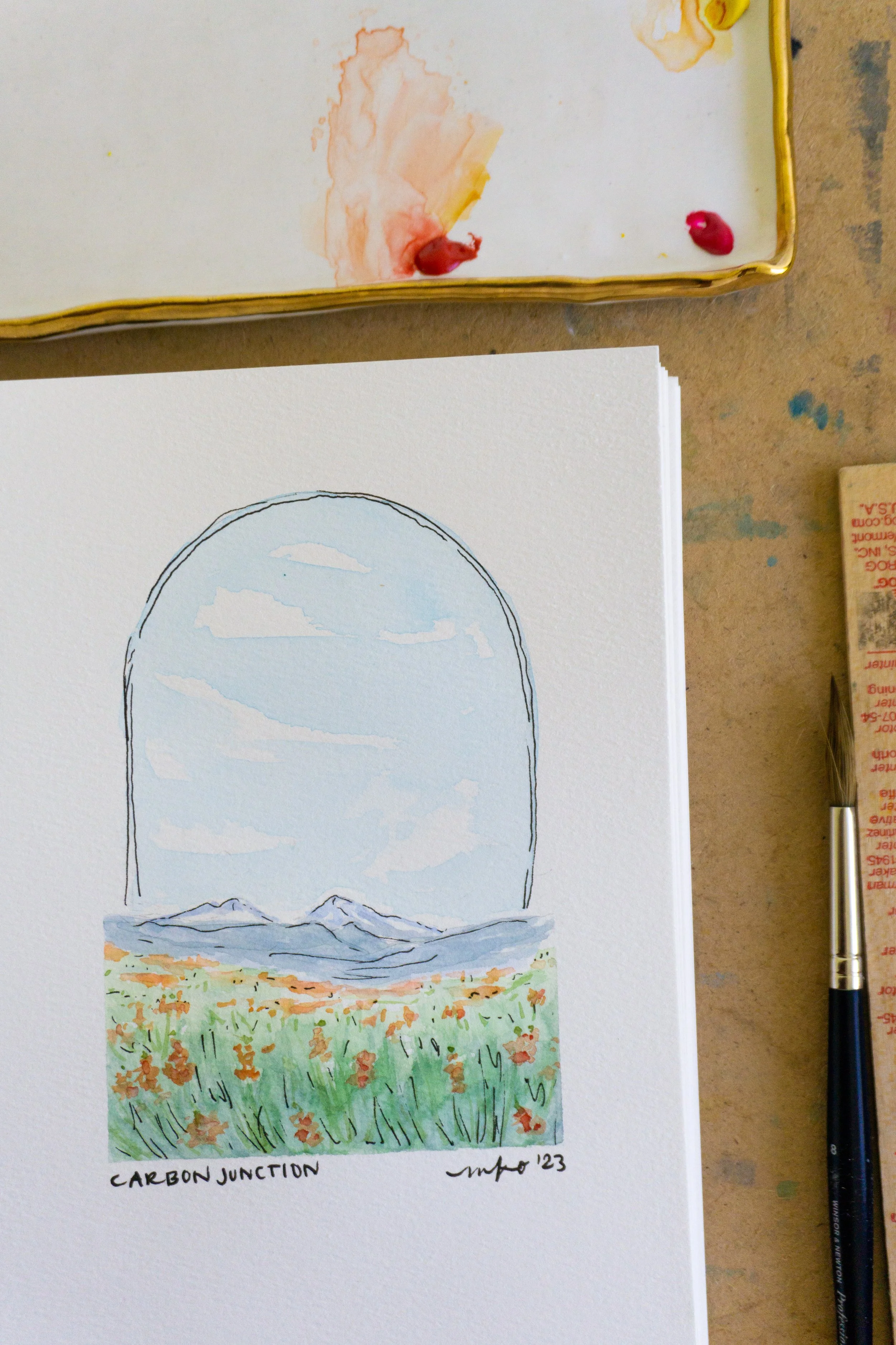 Watercolor painting of a landscape within an arched window frame, depicting mountains, sky, and a field with orange flowers; accompanied by paintbrush and palette.