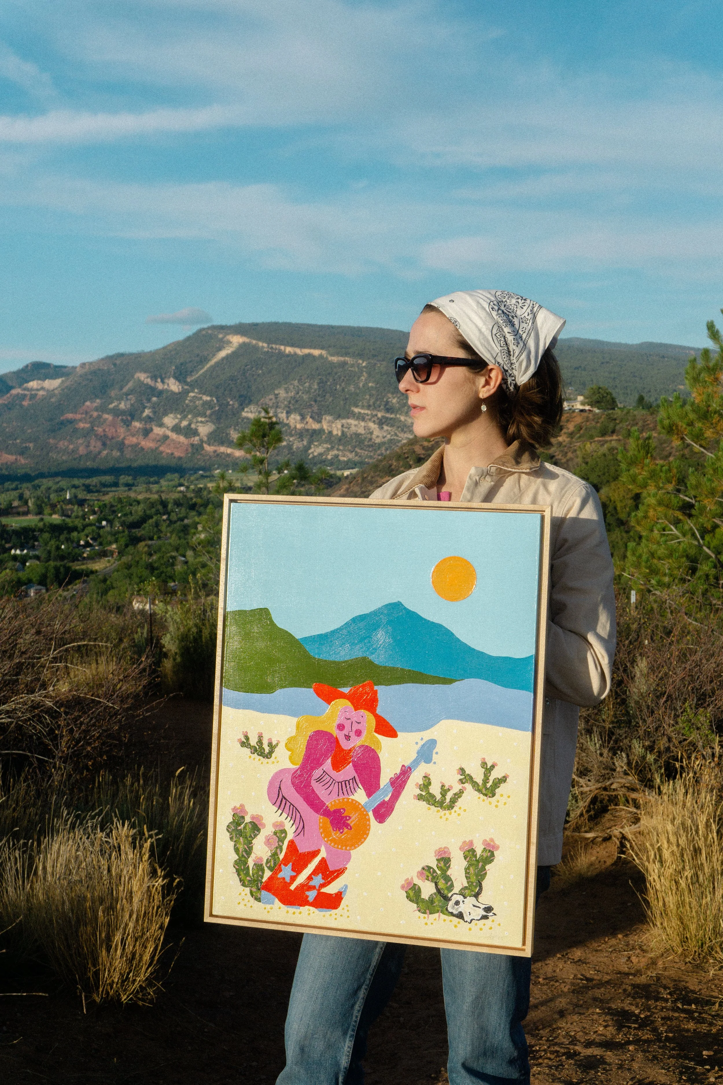 A woman wearing sunglasses, a bandana, and a beige jacket holds a colorful painting of a woman playing a guitar in the desert, with mountains and a blue sky in the background.