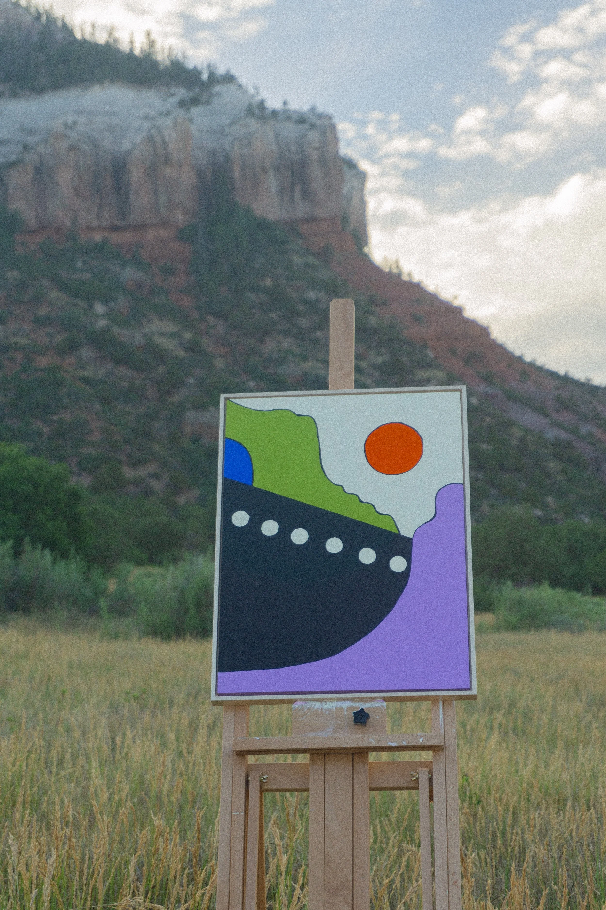 A sign with colorful abstract shapes stands on a wooden easel in a grassy field, with mountains and a partly cloudy sky in the background.