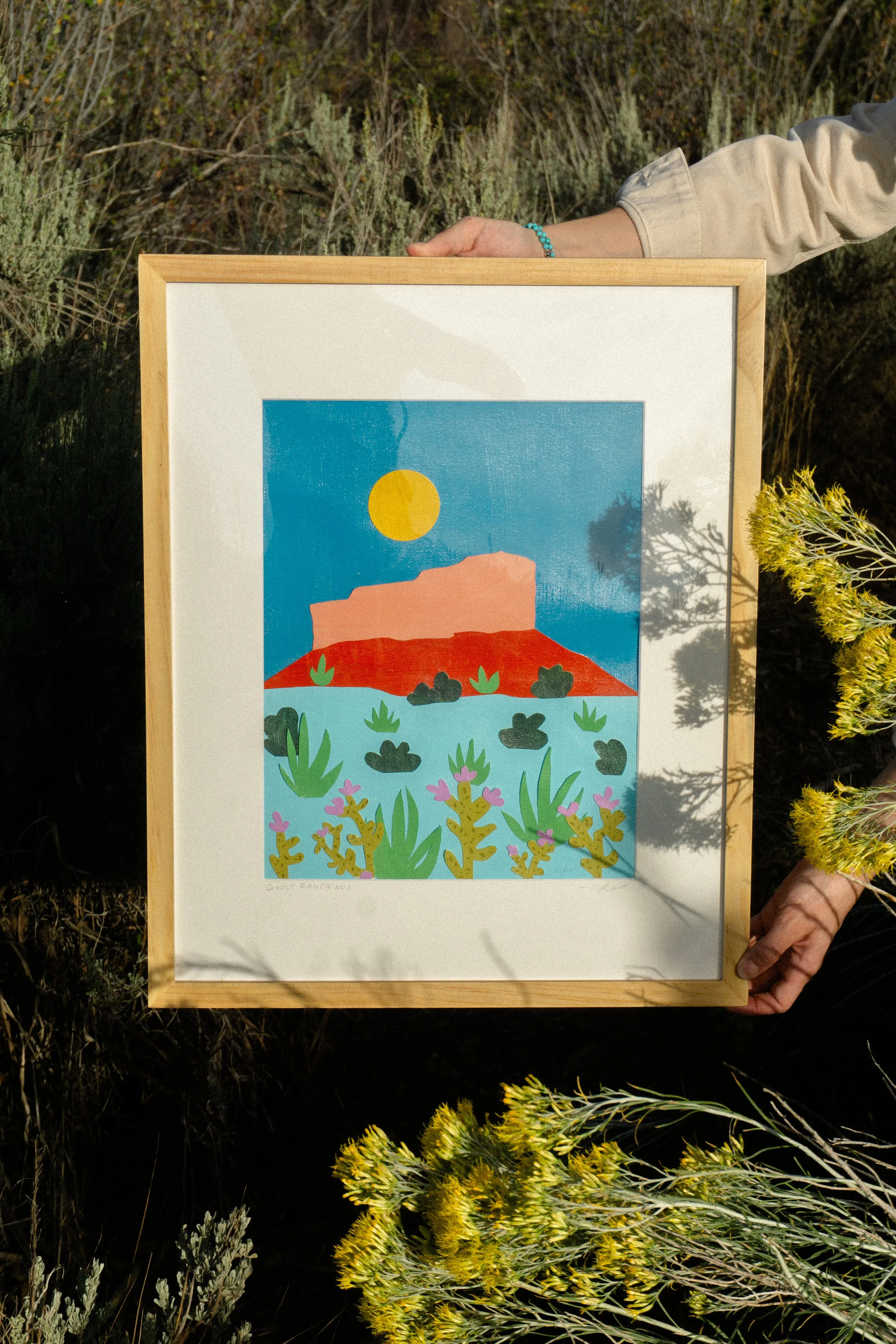 A person holding a framed colorful illustration of a desert landscape with a red butte, blue sky, sun, and various desert plants, outdoors among desert shrubs.
