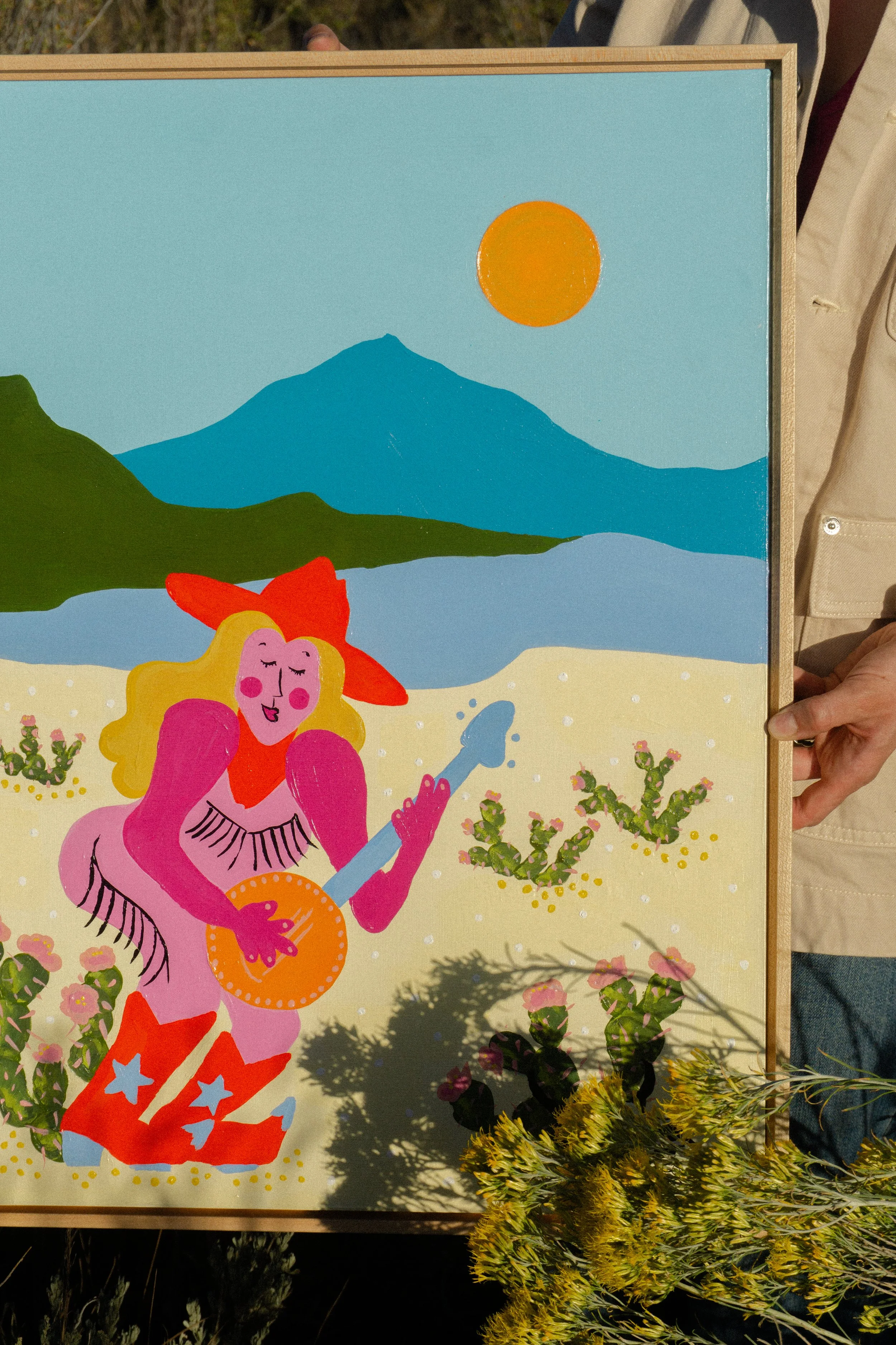 A colorful illustration of a blonde woman with pink skin, wearing a red hat and cowboy boots with  stars, playing a banjo in the desert. In the background, there are mountains, cactuses, and a bright orange sun in the sky.