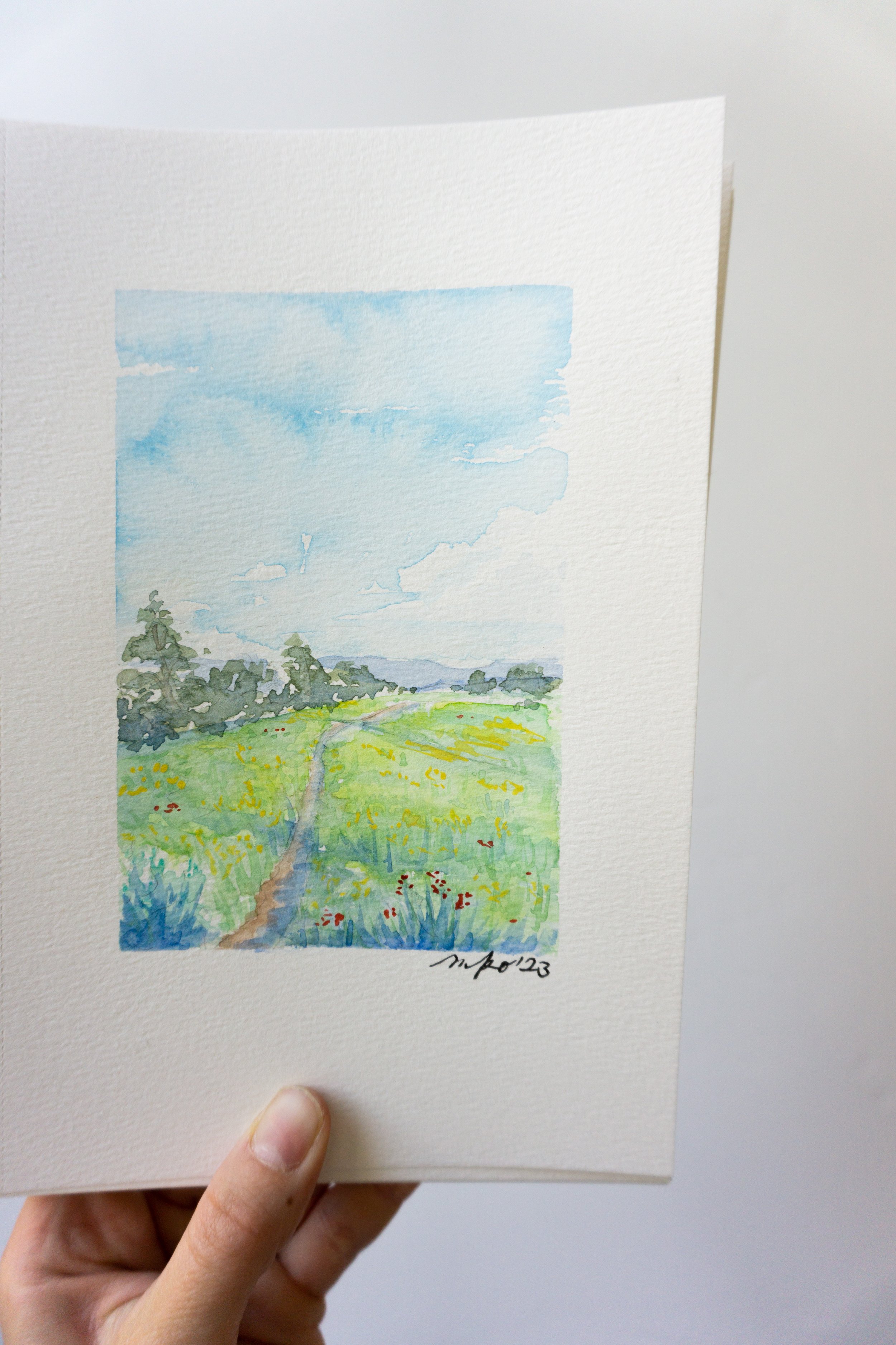 Hand holding a watercolor painting of a landscape with a path through a field, trees, and a cloudy sky.