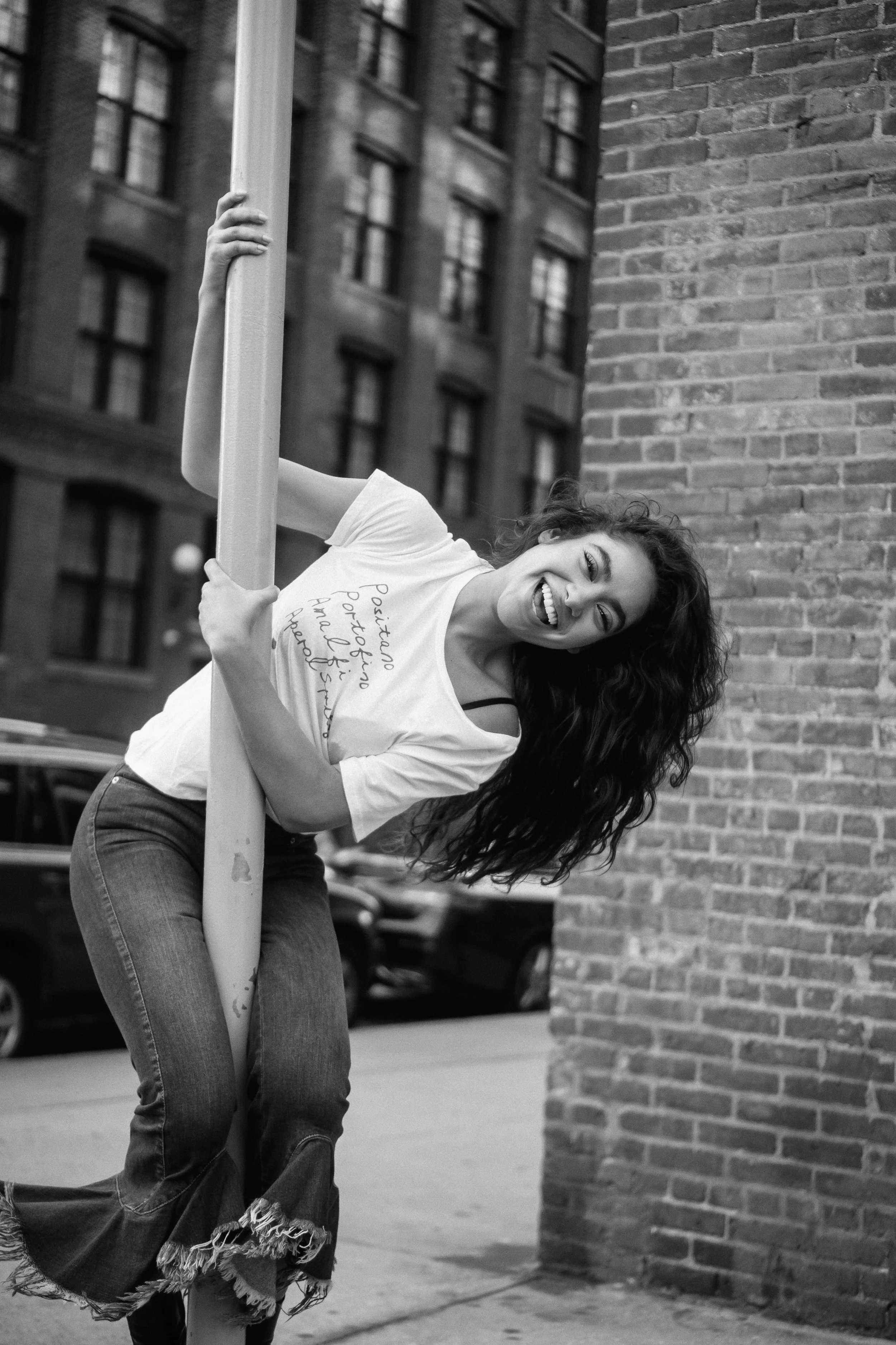 A woman with curly hair smiling and sticking out her tongue, leaning on a pole in an urban setting with brick walls and tall buildings in the background.