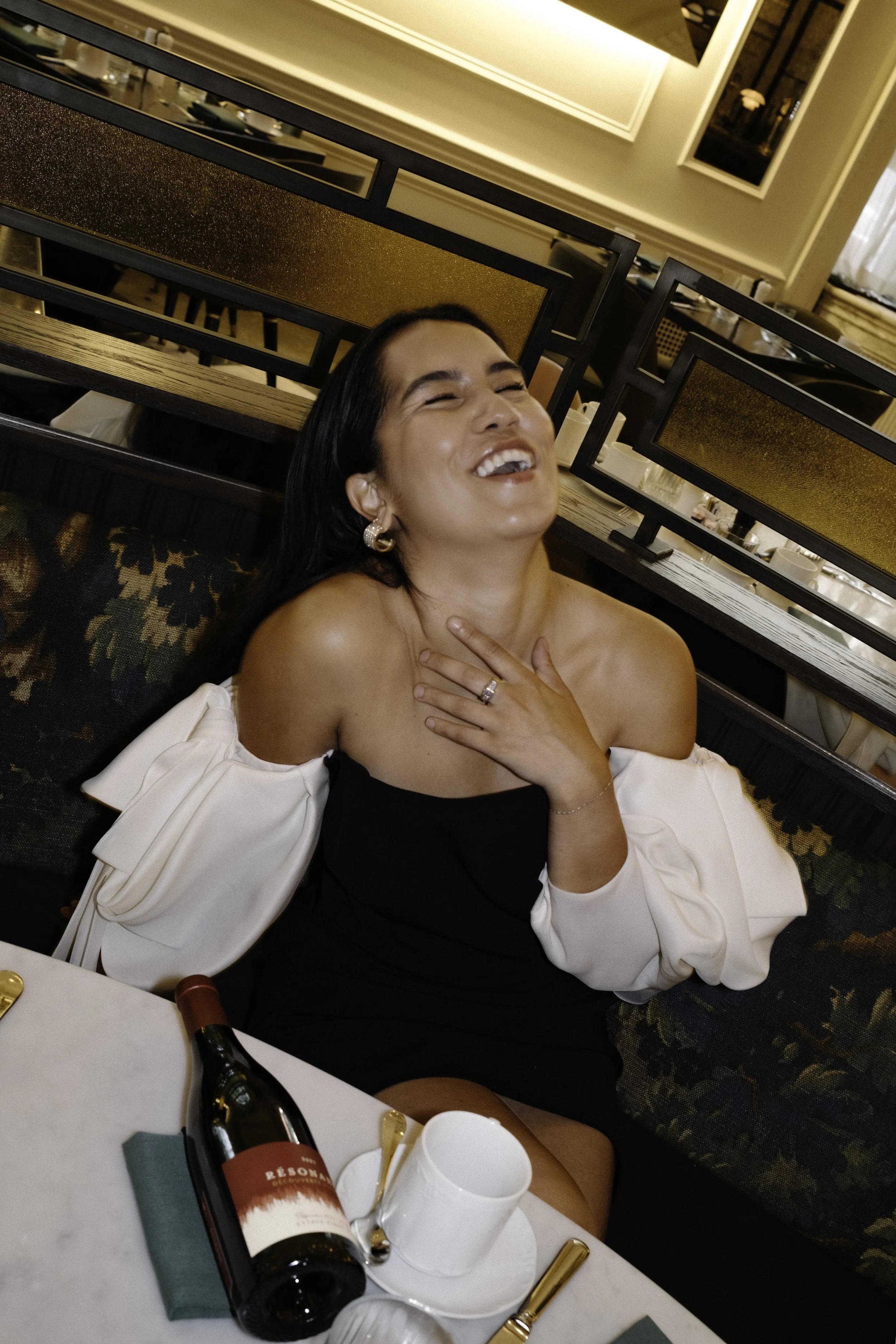 A woman laughing and smiling with her hand on her chest, seated at a restaurant table with a bottle of wine and white teacup in front of her.