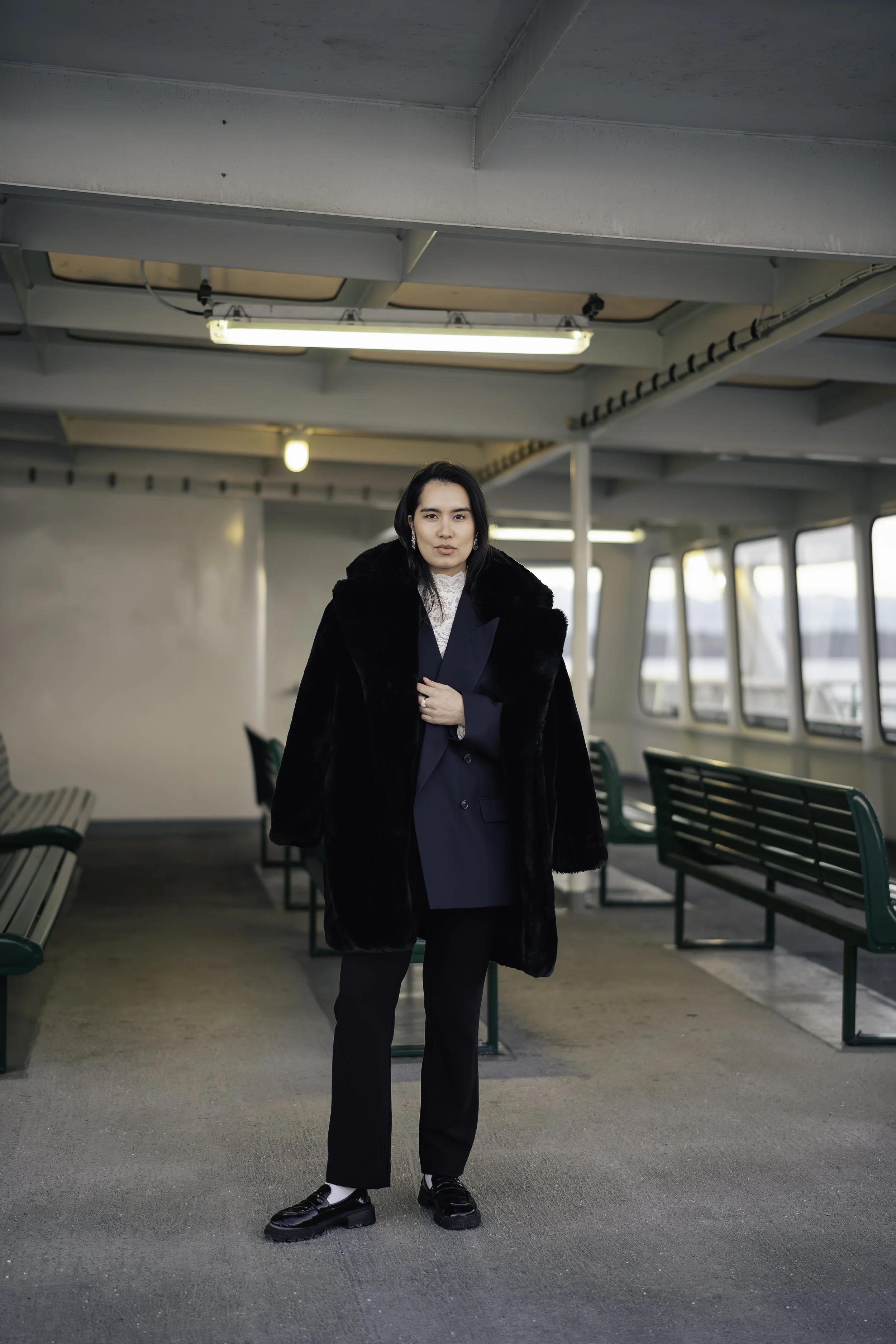 Woman in black fur coat and navy blazer standing inside a boat or ferry with green benches and windows showing water outside.