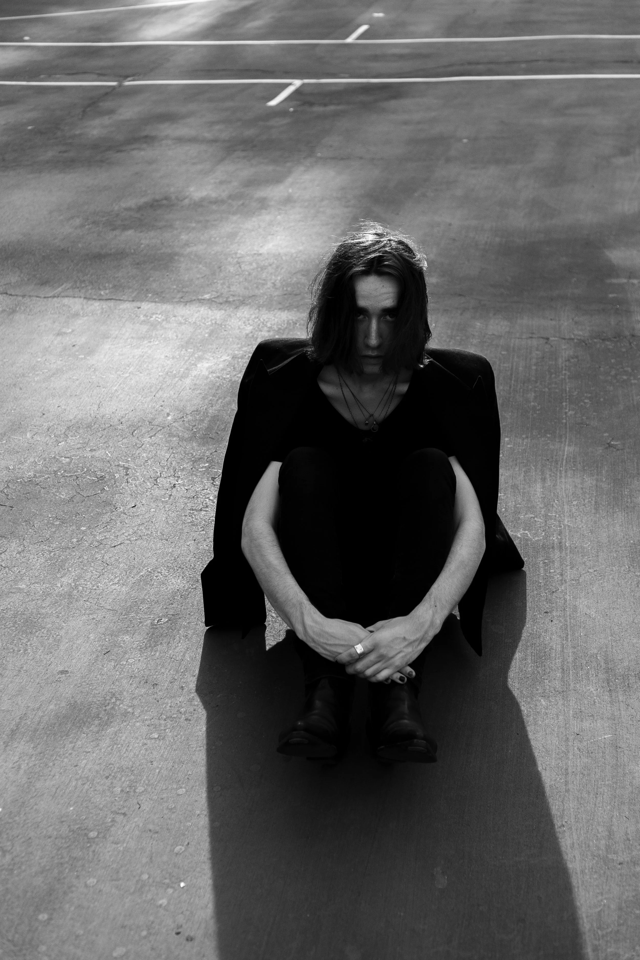 A woman sitting on the ground in an empty parking lot, looking directly at the camera. The photo is in black and white.