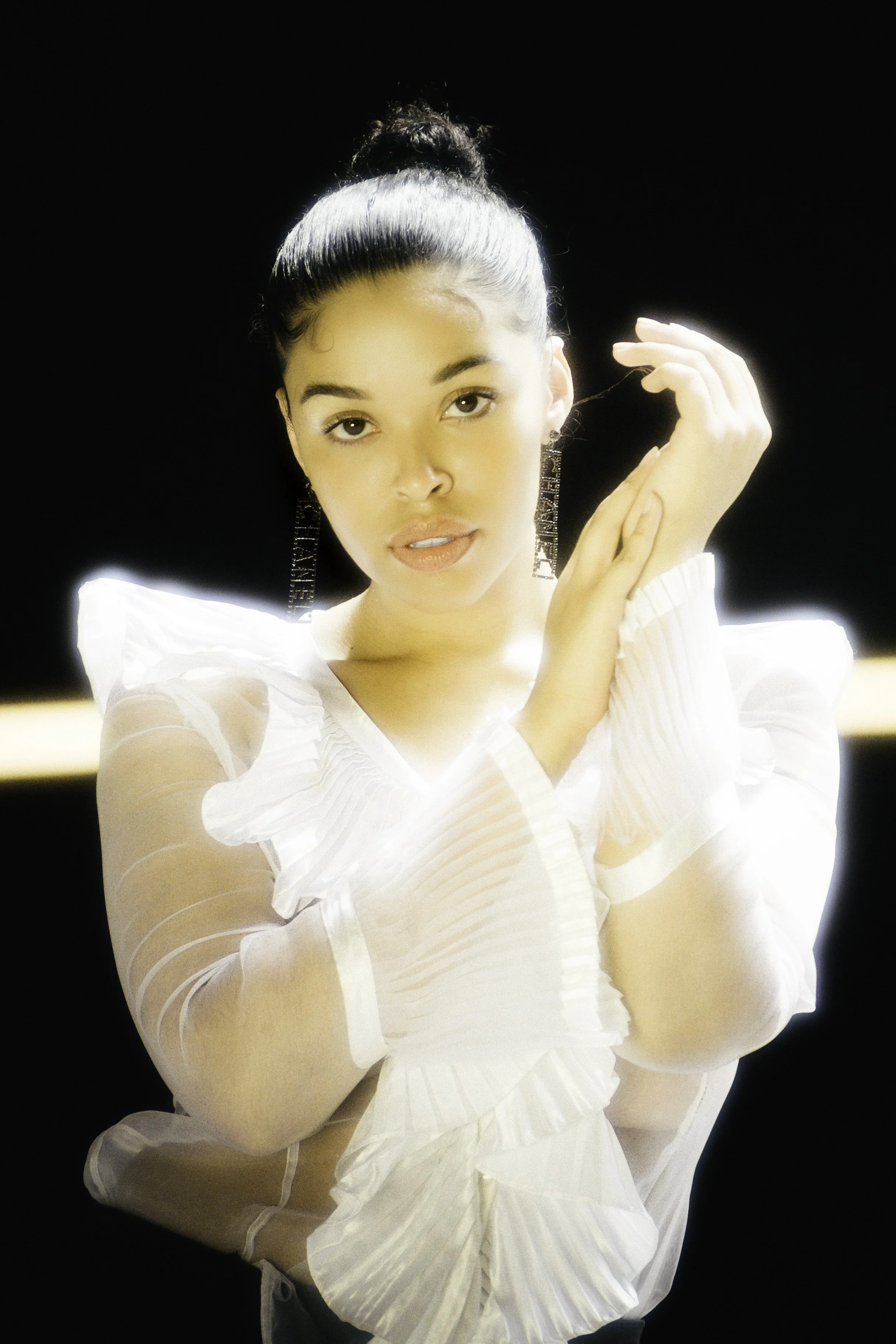 A young woman with dark hair tied up, wearing a white ruffled blouse and long earrings, poses with one hand near her face against a black background with bright lighting.