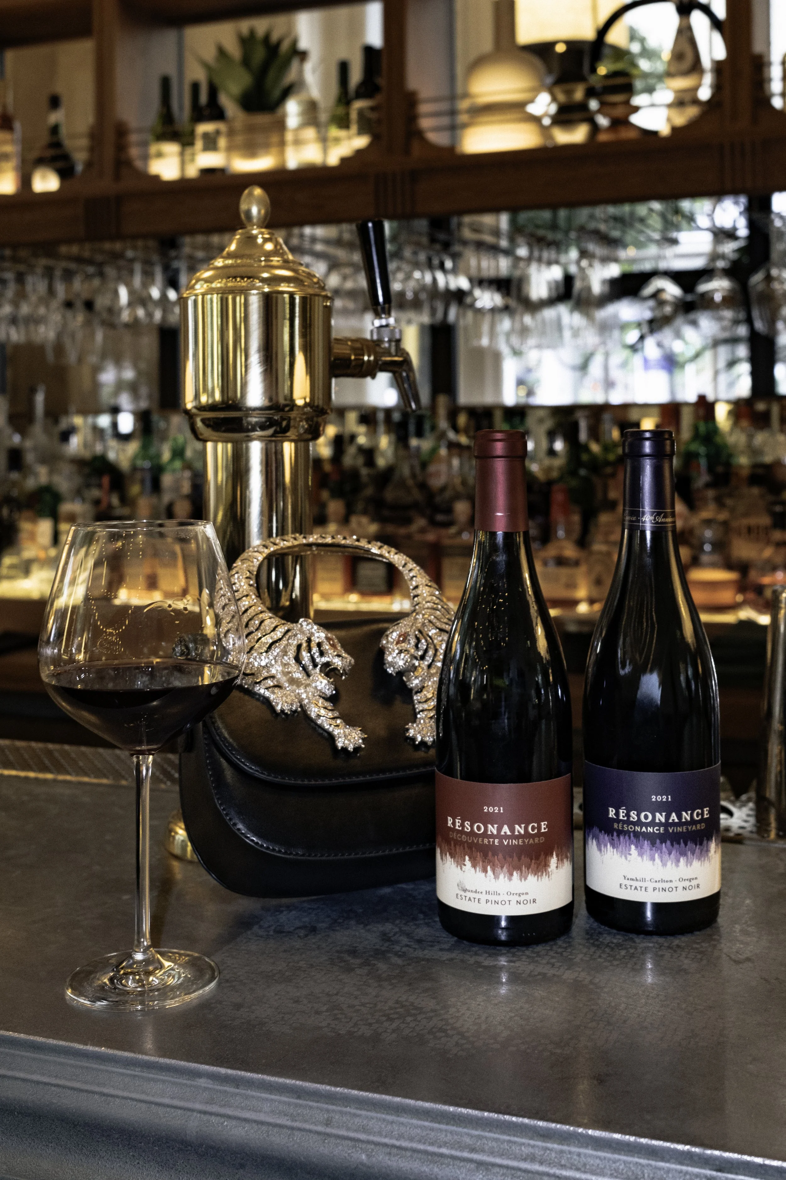 A glass of red wine, two bottles of Resonance Vineyard Pinot Noir wine, a black purse with jeweled tiger head decorations, a brass beer tap, and a bar counter in a restaurant or bar setting.