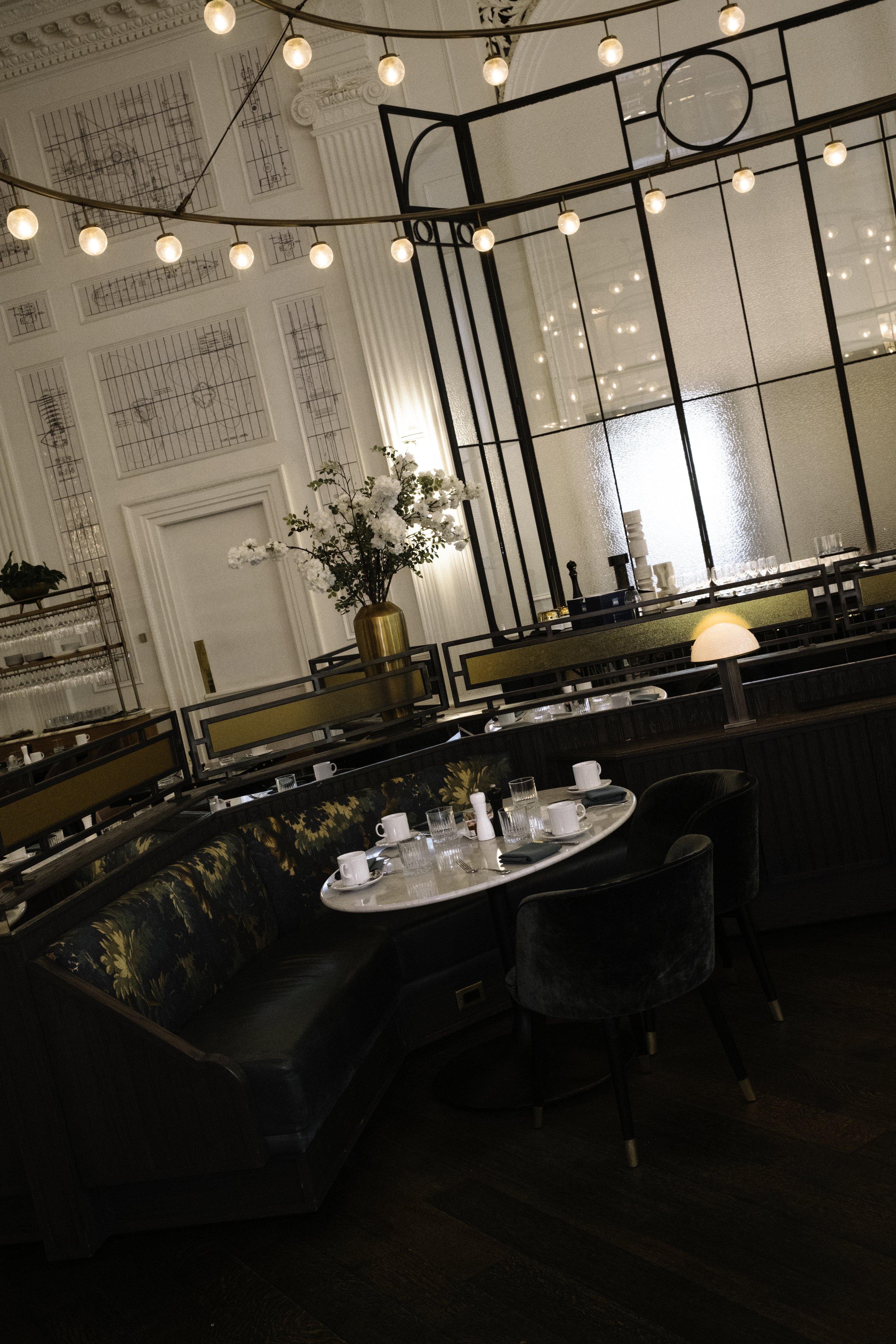 Elegant restaurant interior with a round dining table set with mugs, glasses, and napkins, surrounded by a curved black velvet booth and a dark green velvet chair. There is a large floral arrangement with white flowers in a gold vase, decorative lighting, and modern architectural details in the background.