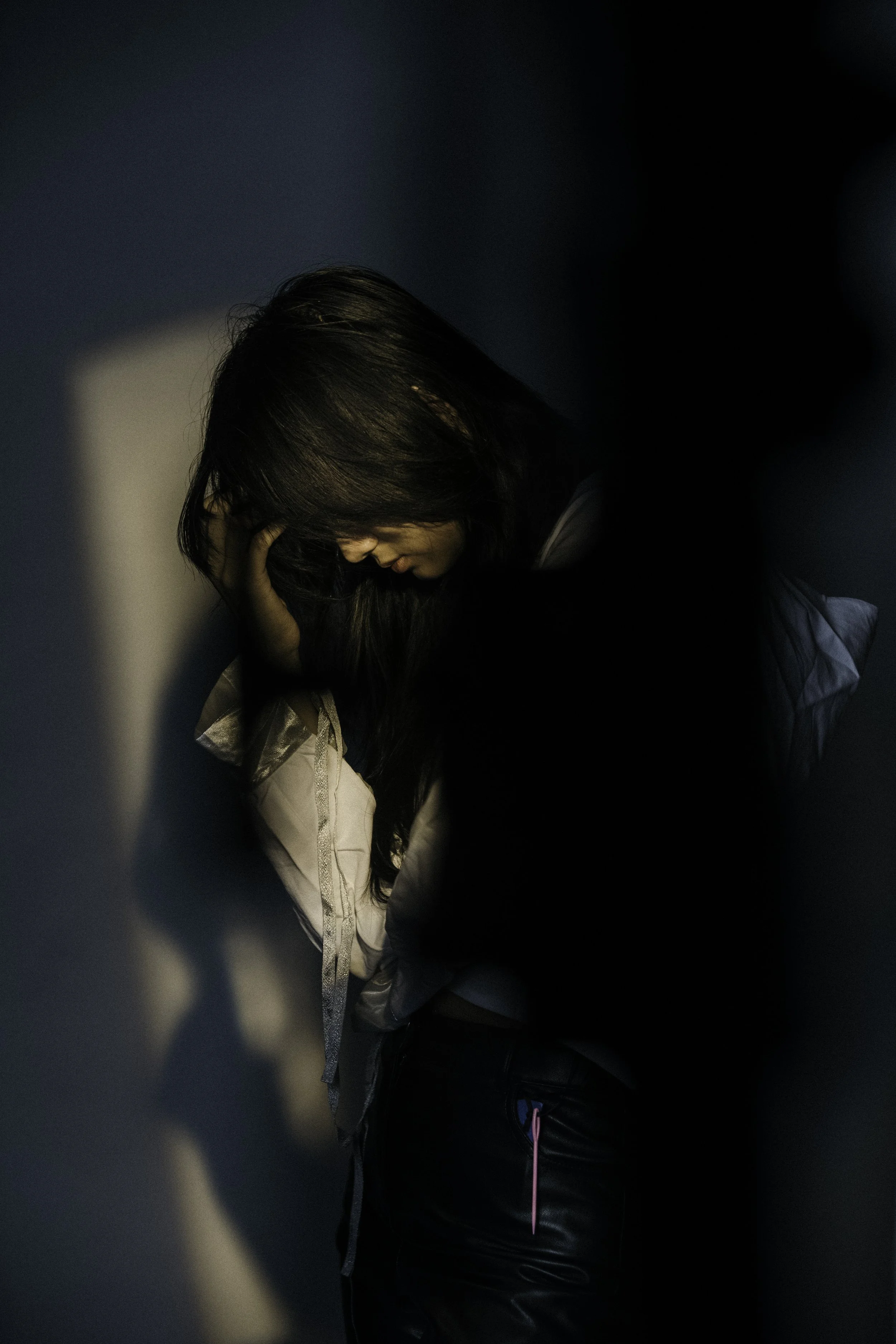 A woman with long dark hair is hugging a child, her face resting against the child's head, in a moment of comfort or affection, with shadows cast on the wall beside them.