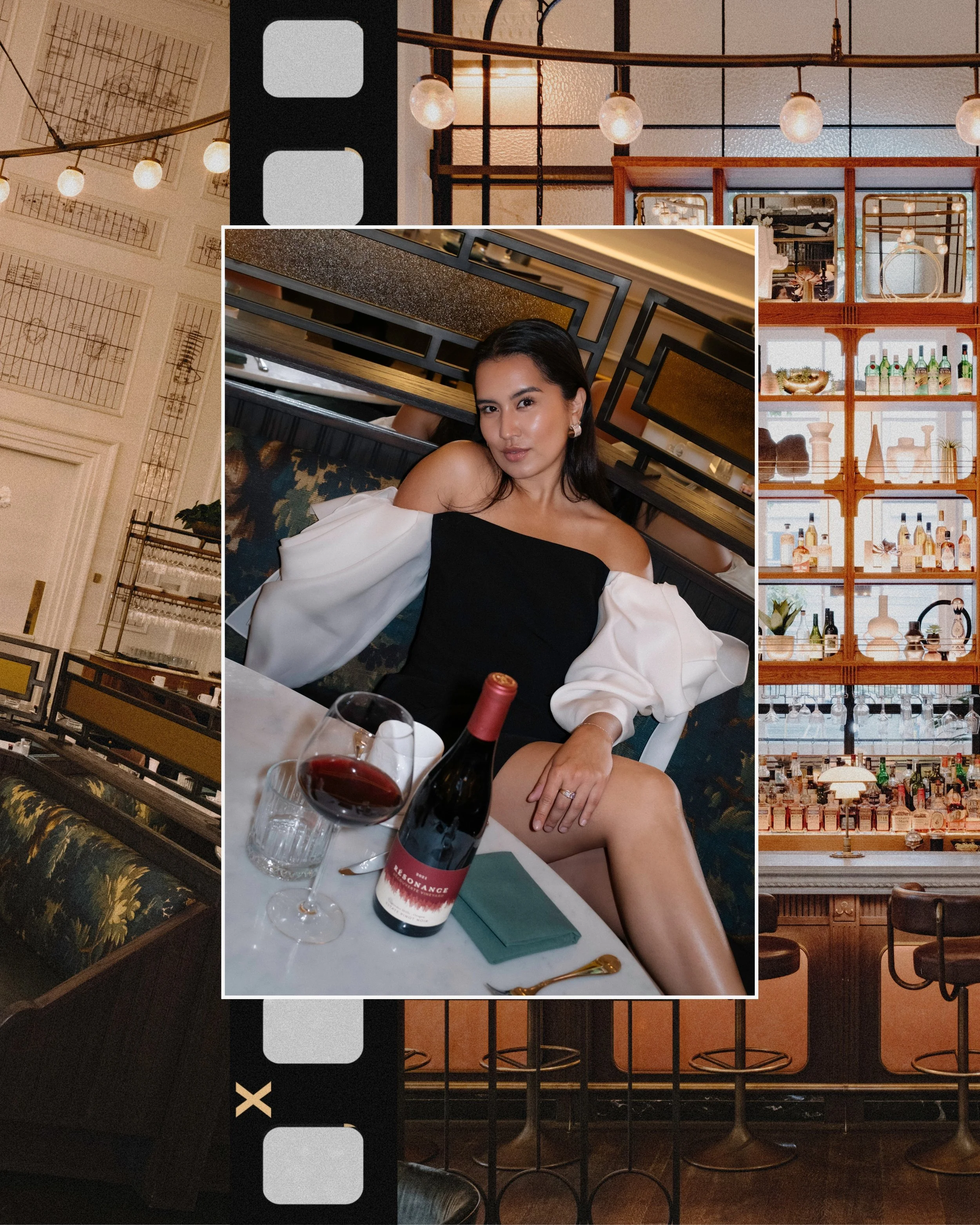 A woman sitting at a restaurant table with a glass of red wine and a bottle of wine in front of her, wearing a black and white off-shoulder dress, in an upscale bar or restaurant with elegant decor and warm lighting.