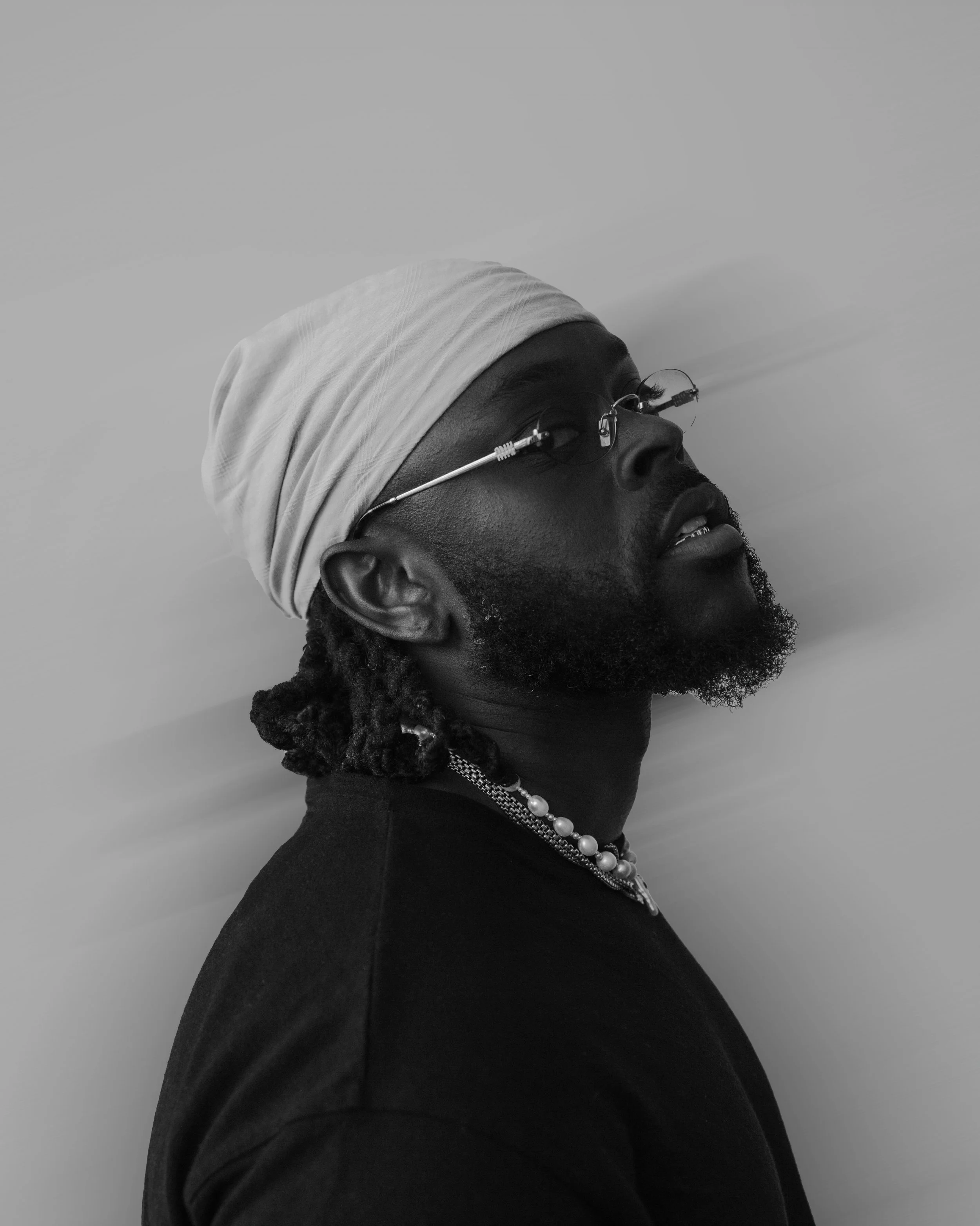 A black and white portrait of a man with dreadlocks wearing a head wrap, glasses, a pearl necklace, and a dark shirt, looking slightly upward.