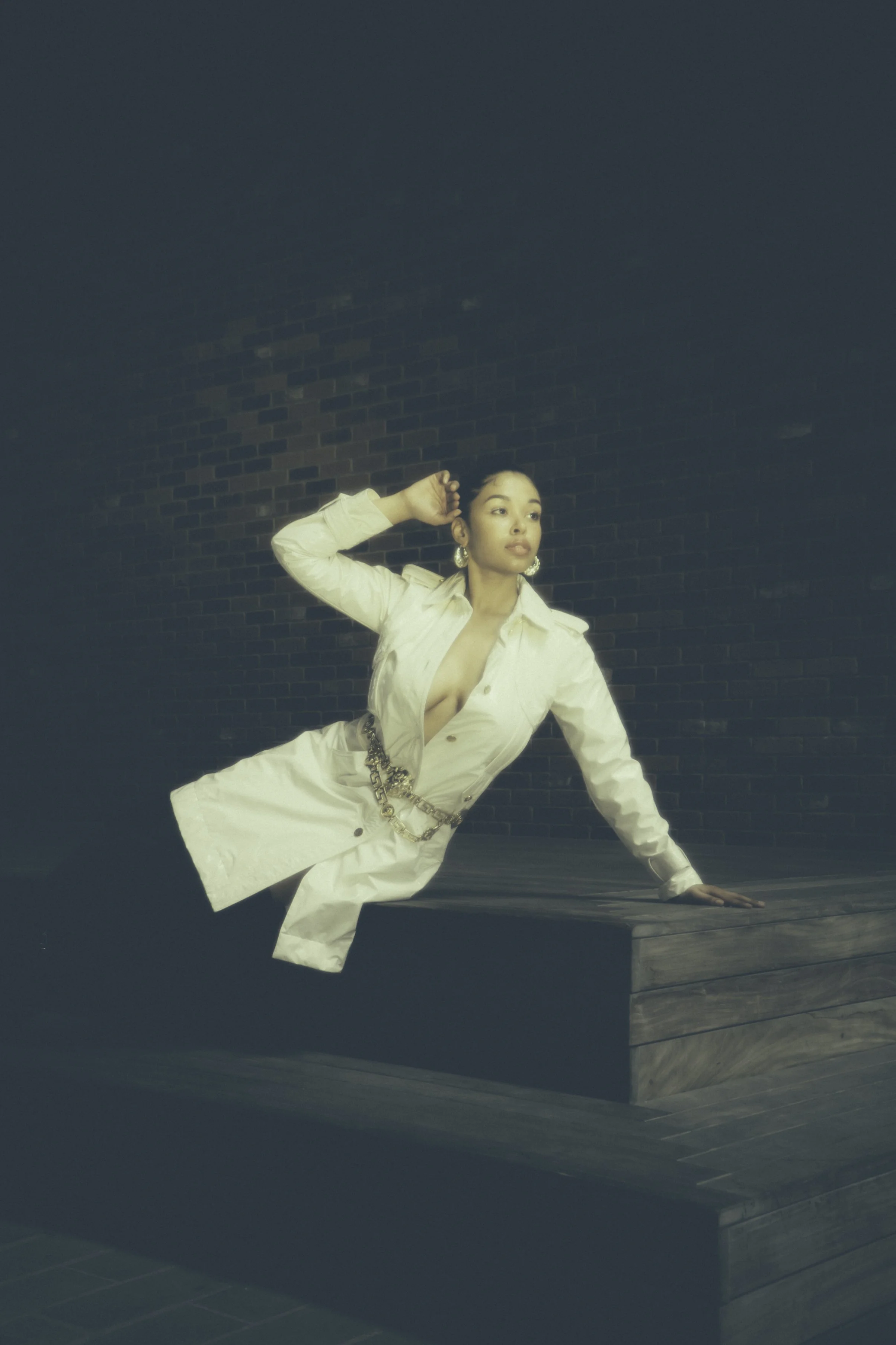 A woman in a beige trench coat with a chain belt poses outdoors against a brick wall, standing on a wooden surface during daytime.