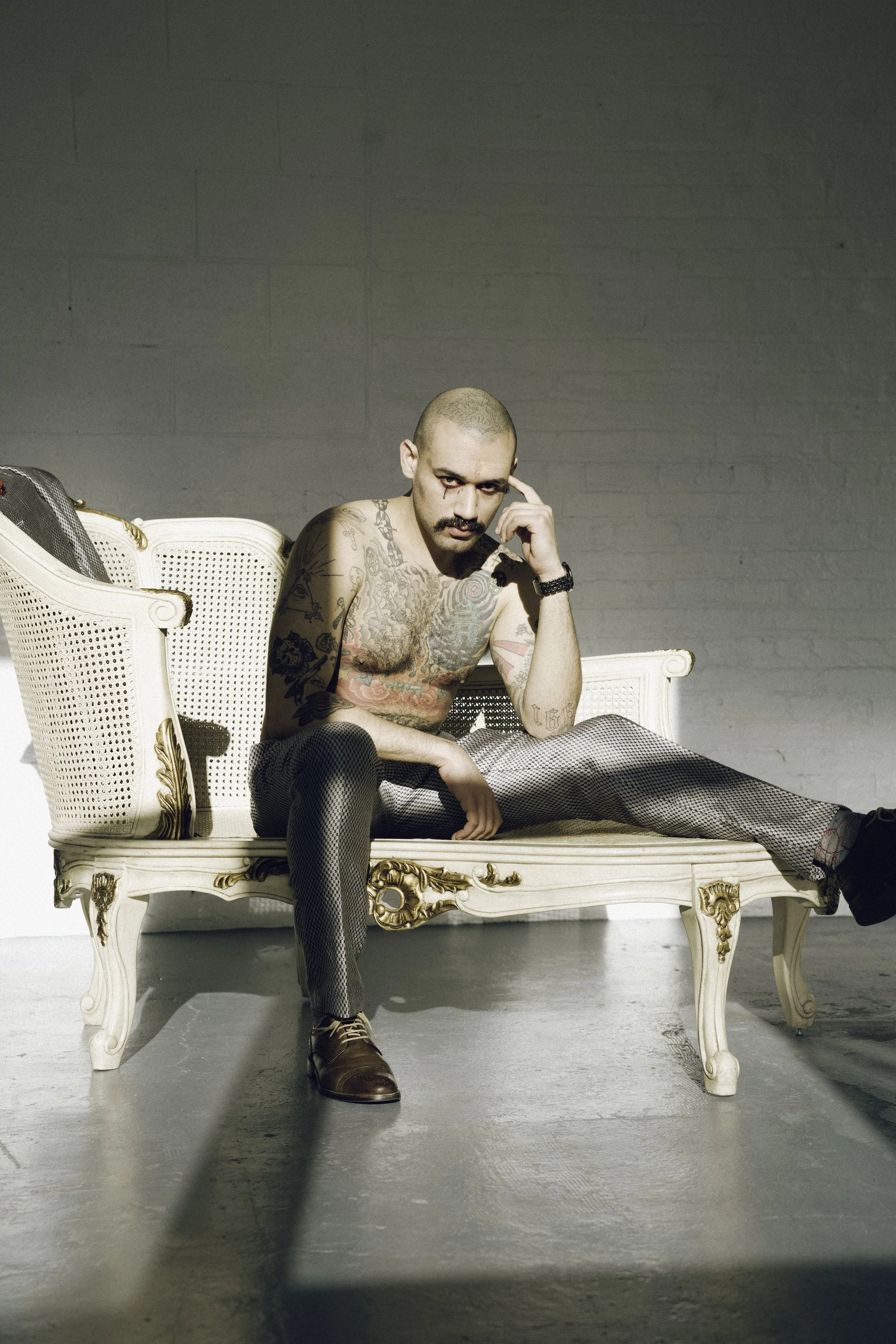 Tattooed man with a shaved head sitting on an ornate vintage sofa against a plain wall, wearing patterned pants, a watch, and boots, with a contemplative expression.