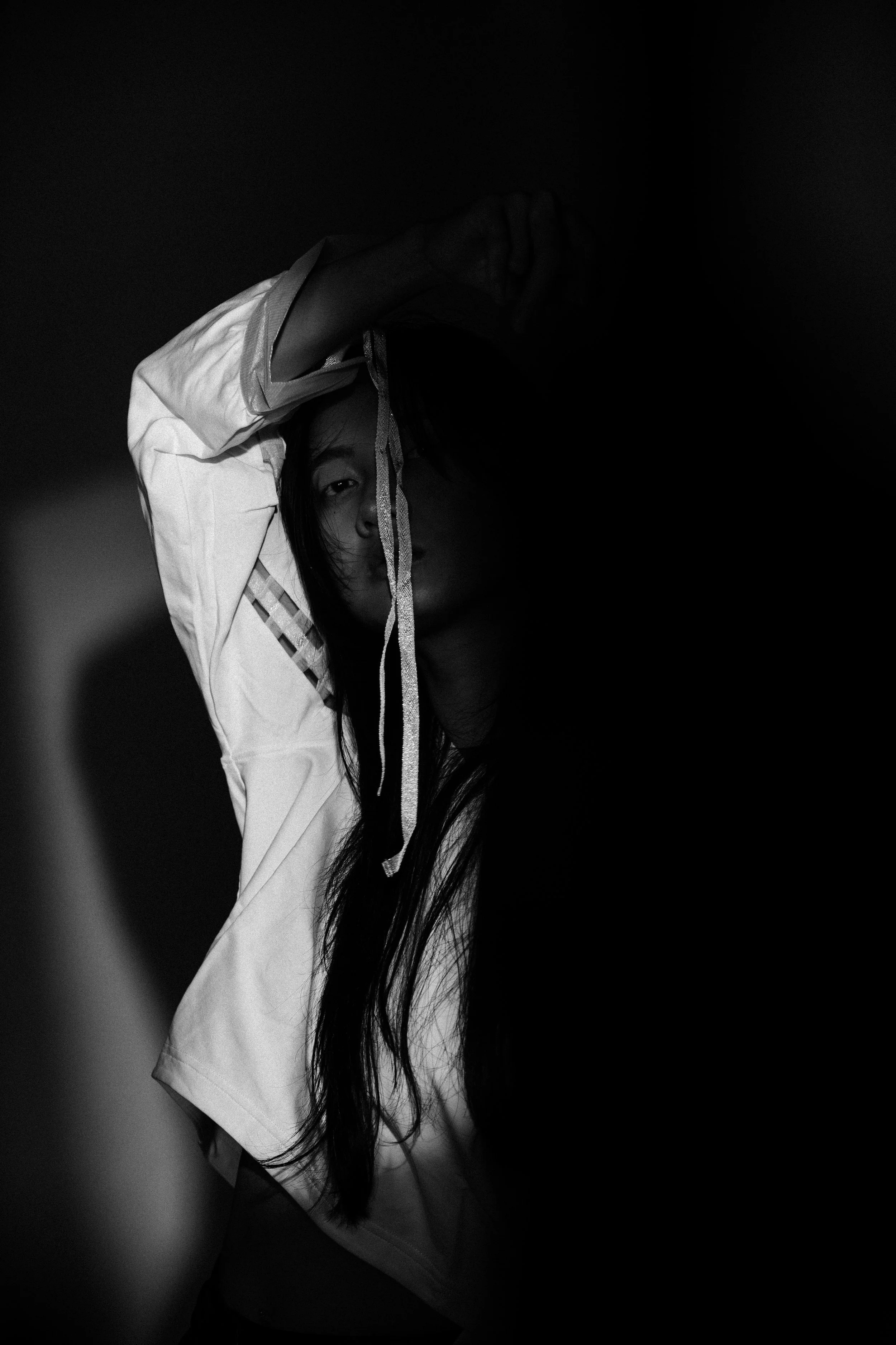 A woman with long dark hair poses with one arm raised above her head, partially covering her face, in a black-and-white photo with dramatic lighting and shadows.